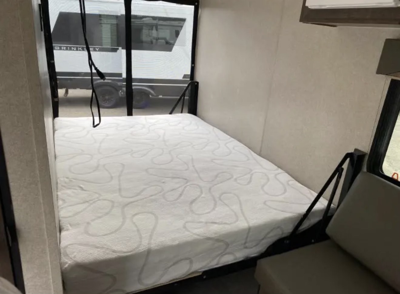 Compact RV rental bedroom with a comfy white patterned mattress. Window view shows another RV outside, next to a seating area.. Sunset Park RV SunRay 2022