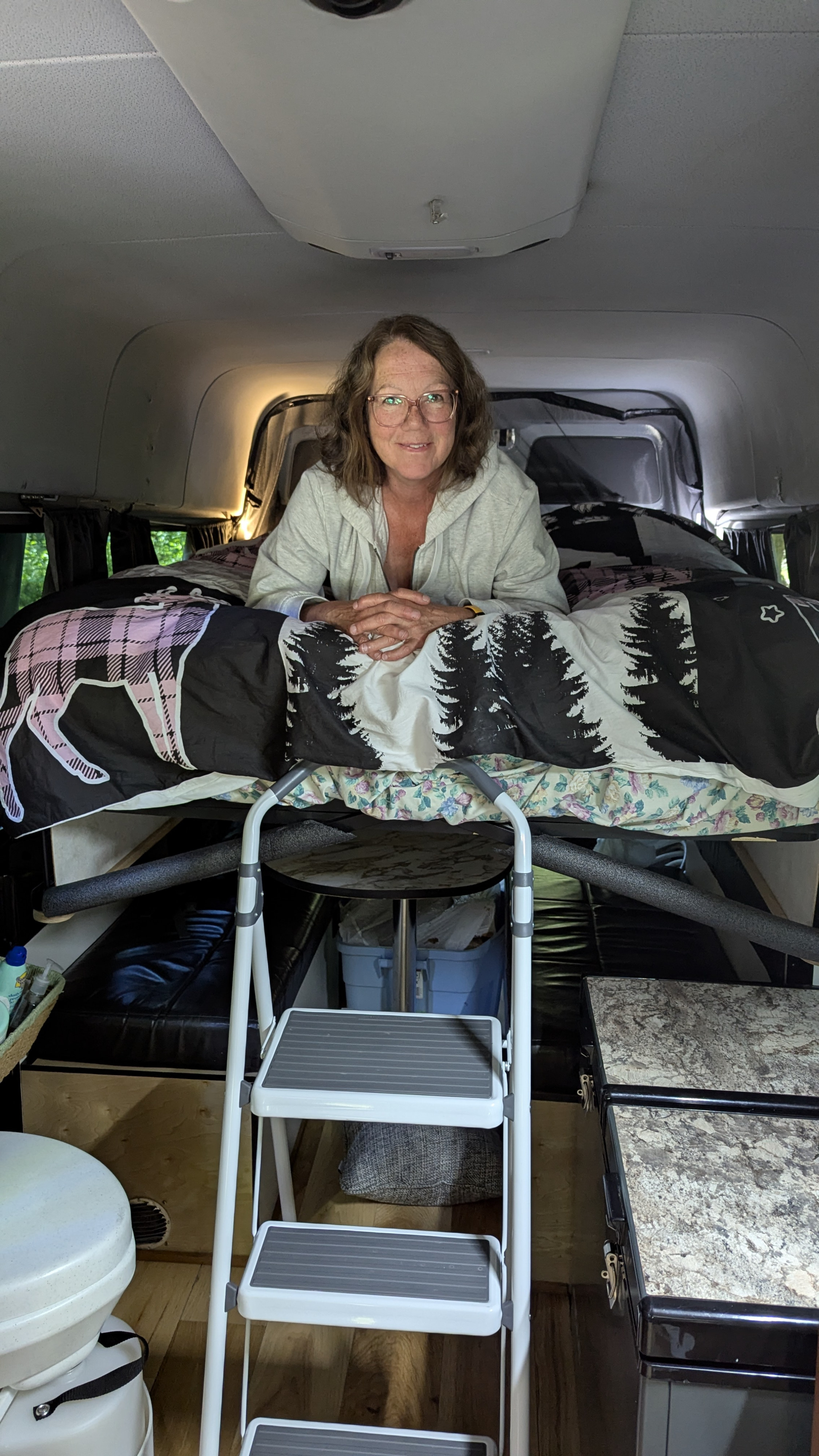 Smiling woman on a loft bed in her camper van, featuring a ladder, toilet, and storage. Ideal for your next RV rental trip!. Mercedes Sprinter 2008