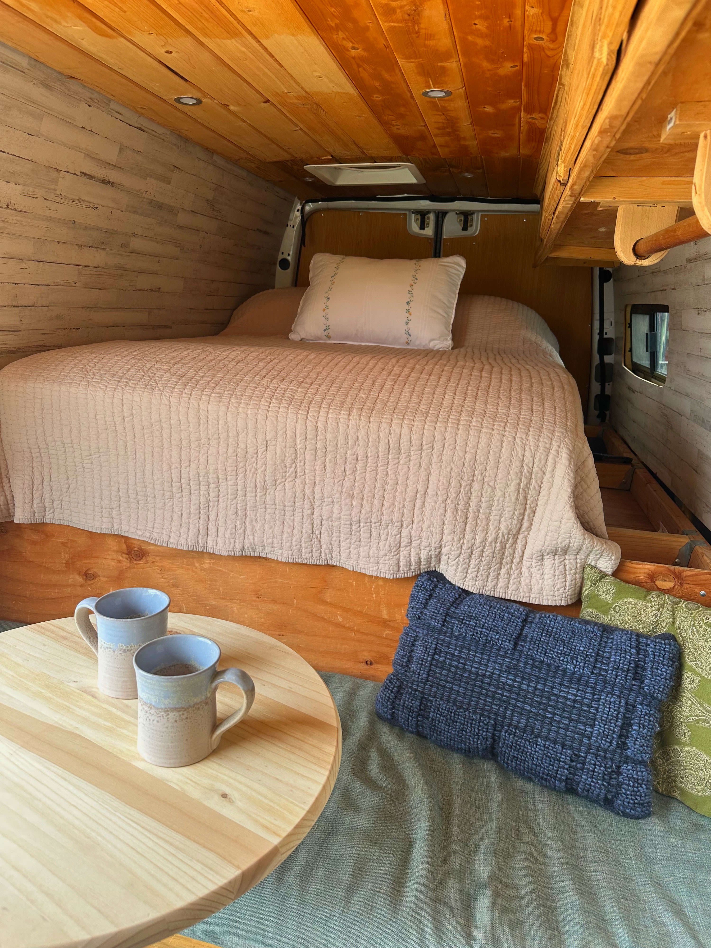 This rv rental shows a cozy van conversion interior. Features a comfy bed, wooden accents, mugs on a table, and soft pillows.. Mercedes-Benz Sprinter 316 2008