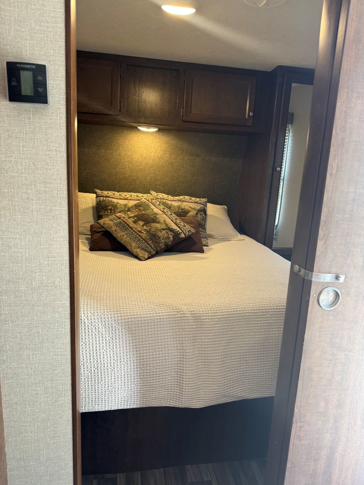 RV rental bedroom with a cozy bed, rustic pillows, overhead storage cabinets, and a thermostat on the wall.. Keystone RV Hideout 2018