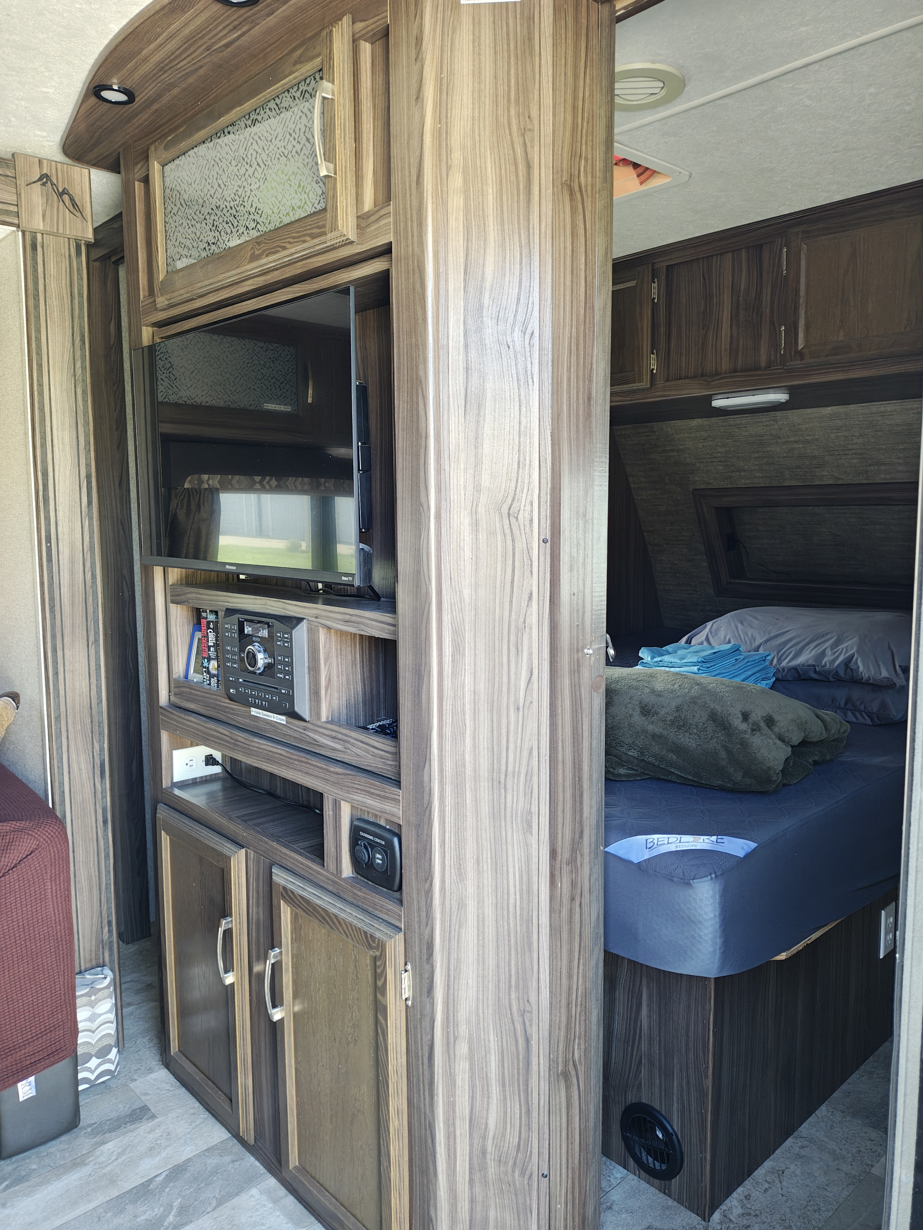 RV rental interior featuring a wood-paneled entertainment center with TV/stereo, storage, and a cozy bedroom with a Bedlore mattress.. Coachmen Apex 2018