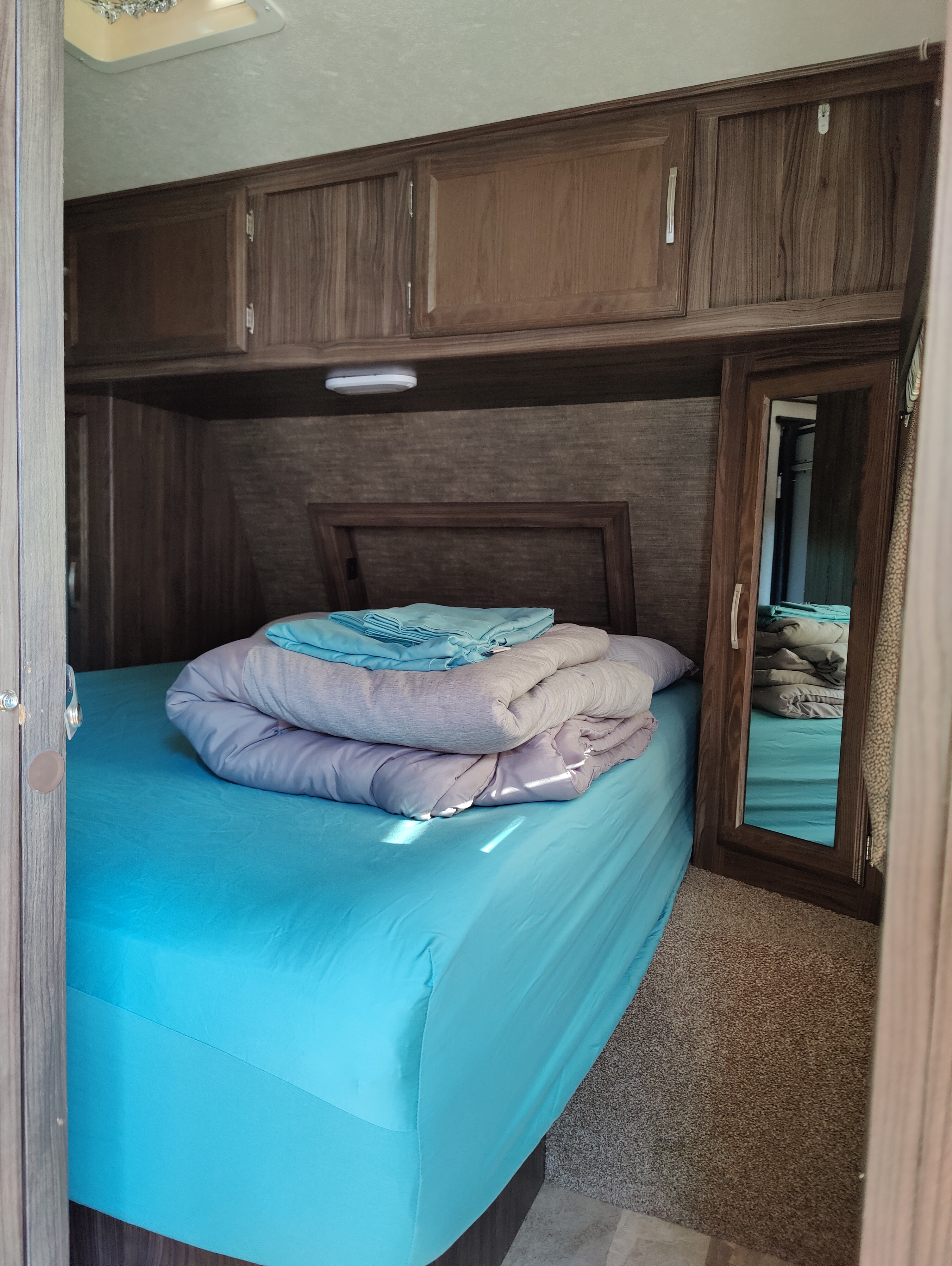 RV rental bedroom with a bright blue bed, neatly folded blankets, overhead wooden cabinets, and a mirrored closet for storage.. Coachmen Apex 2018