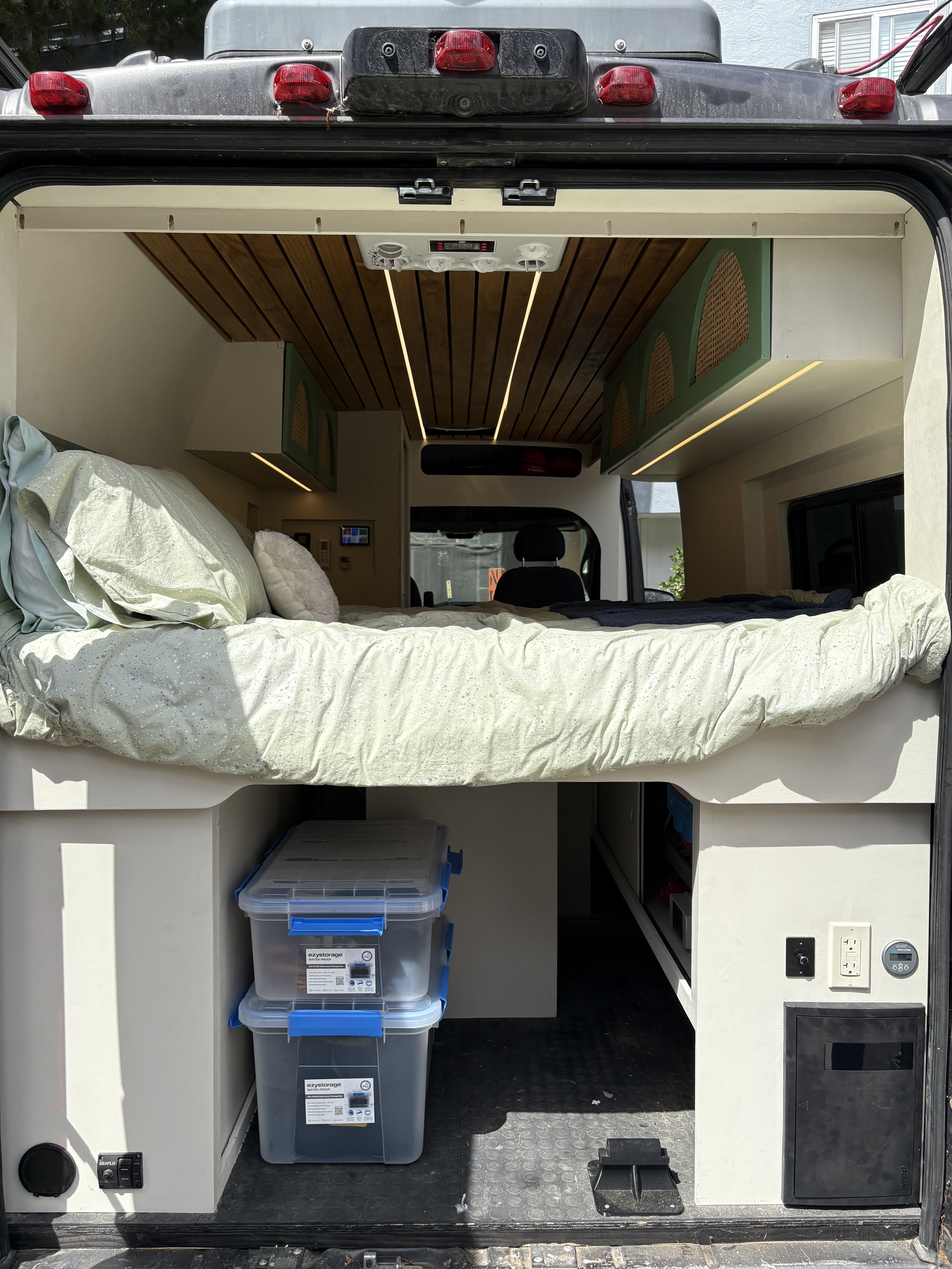 RV rental interior, seen from the rear. A cozy bed is elevated over under-bed storage, electrical controls, and wood-paneled ceiling.. Ram Promaster 2500 2017