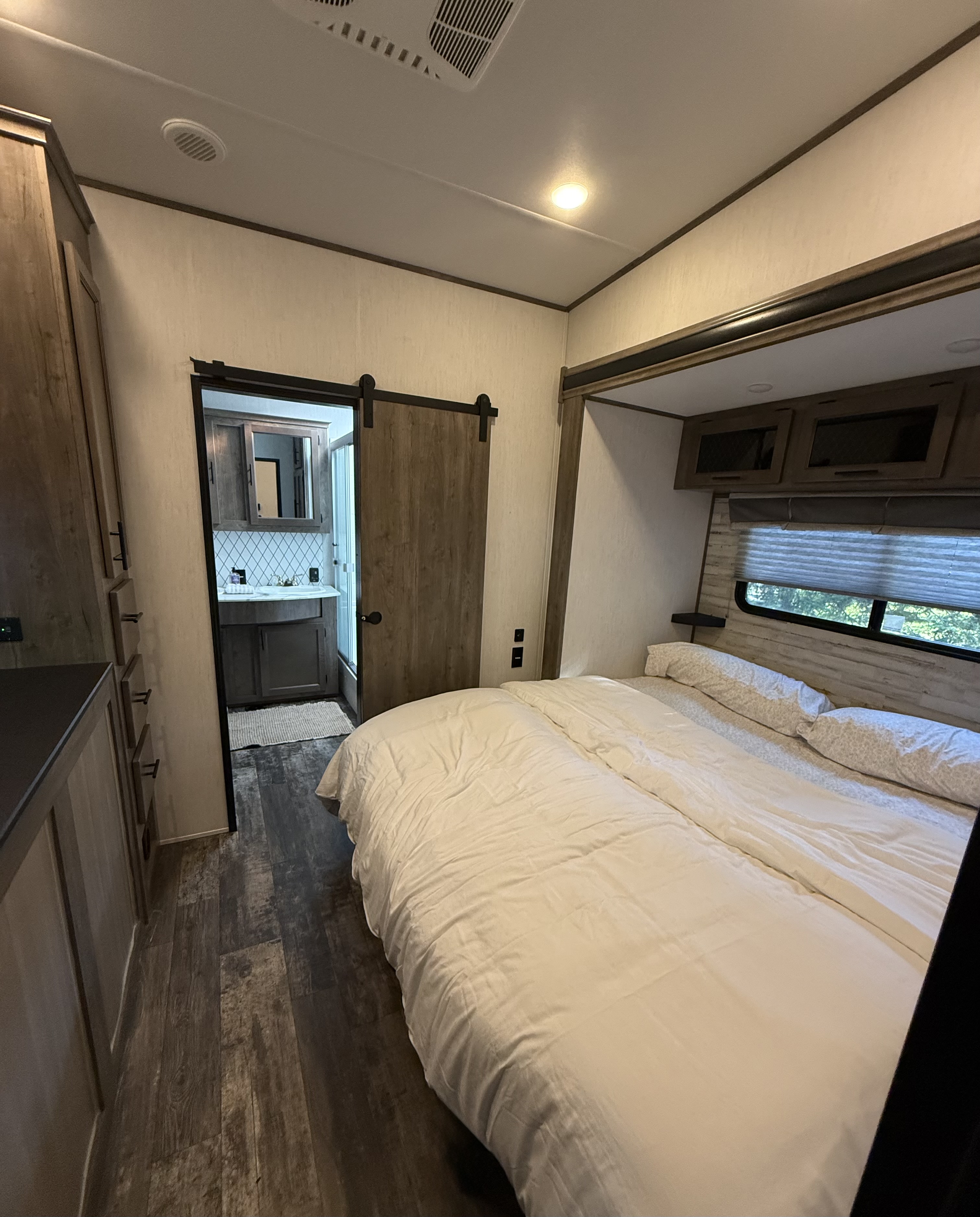 RV rental bedroom: a cozy retreat with a large bed, wood-look floors, overhead storage, and a modern bathroom behind a barn door.. Forest River Sabre 2021