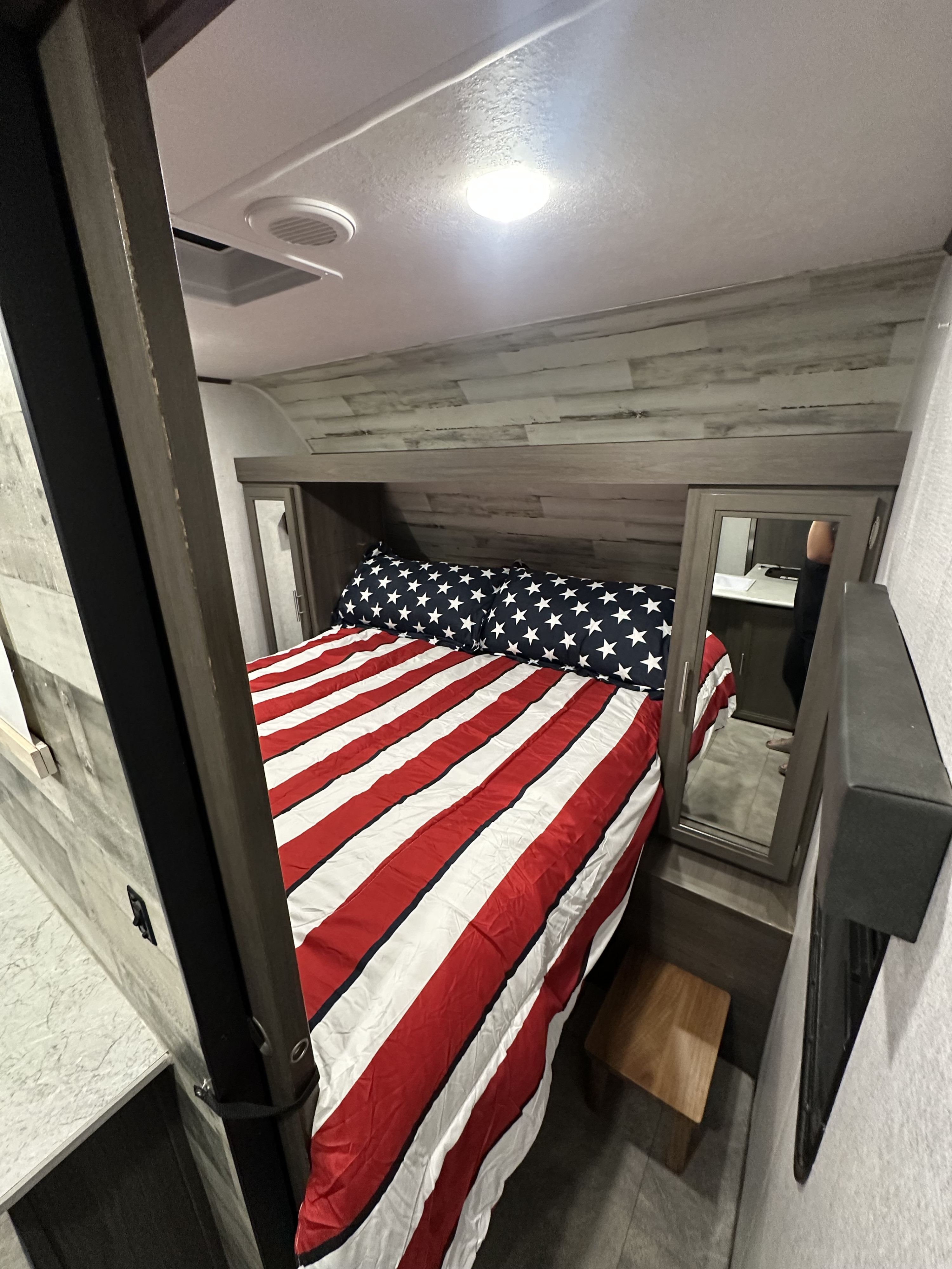 Compact RV rental bedroom with patriotic American flag bedding, ceiling light, air vent, mirrored cabinet, and wood paneling.. Dutchmen Kodiak SE 2021