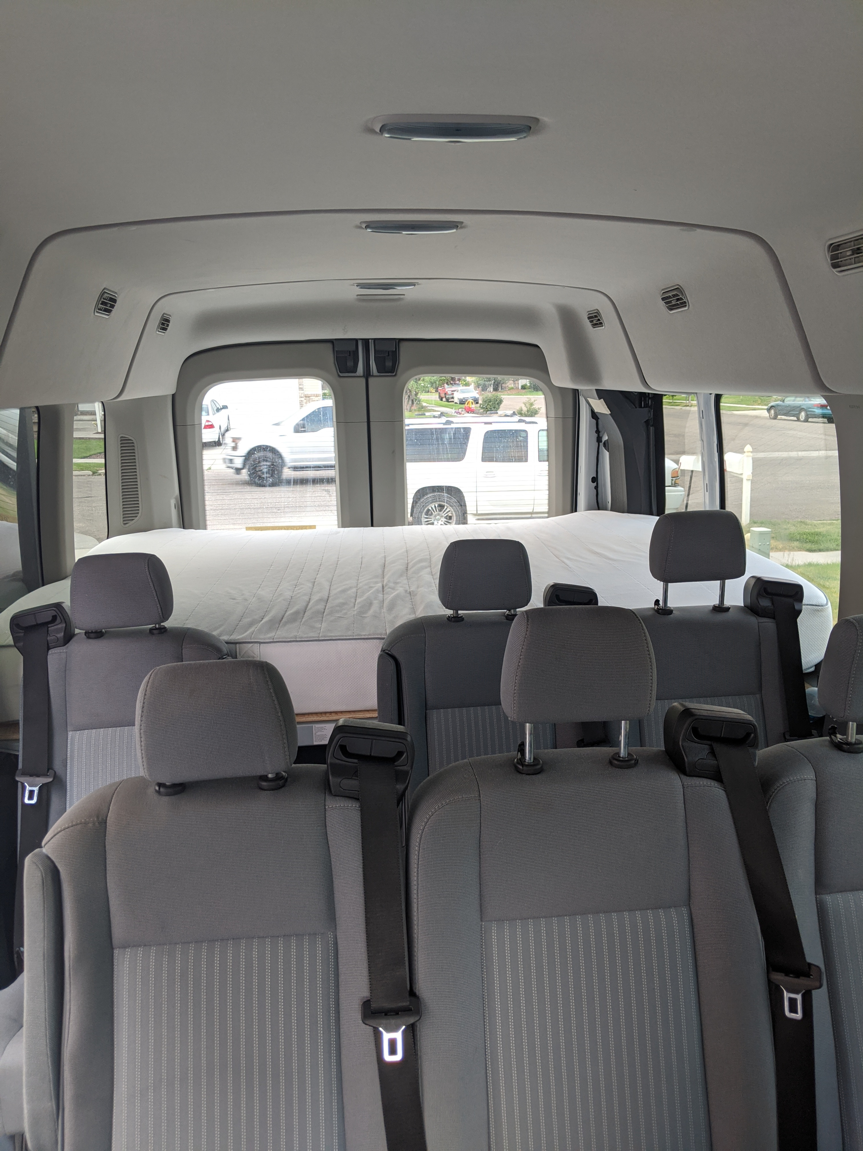 View of a van interior, likely an rv rental, featuring grey passenger seats and a large white mattress in the back.. Ford Transit 2016