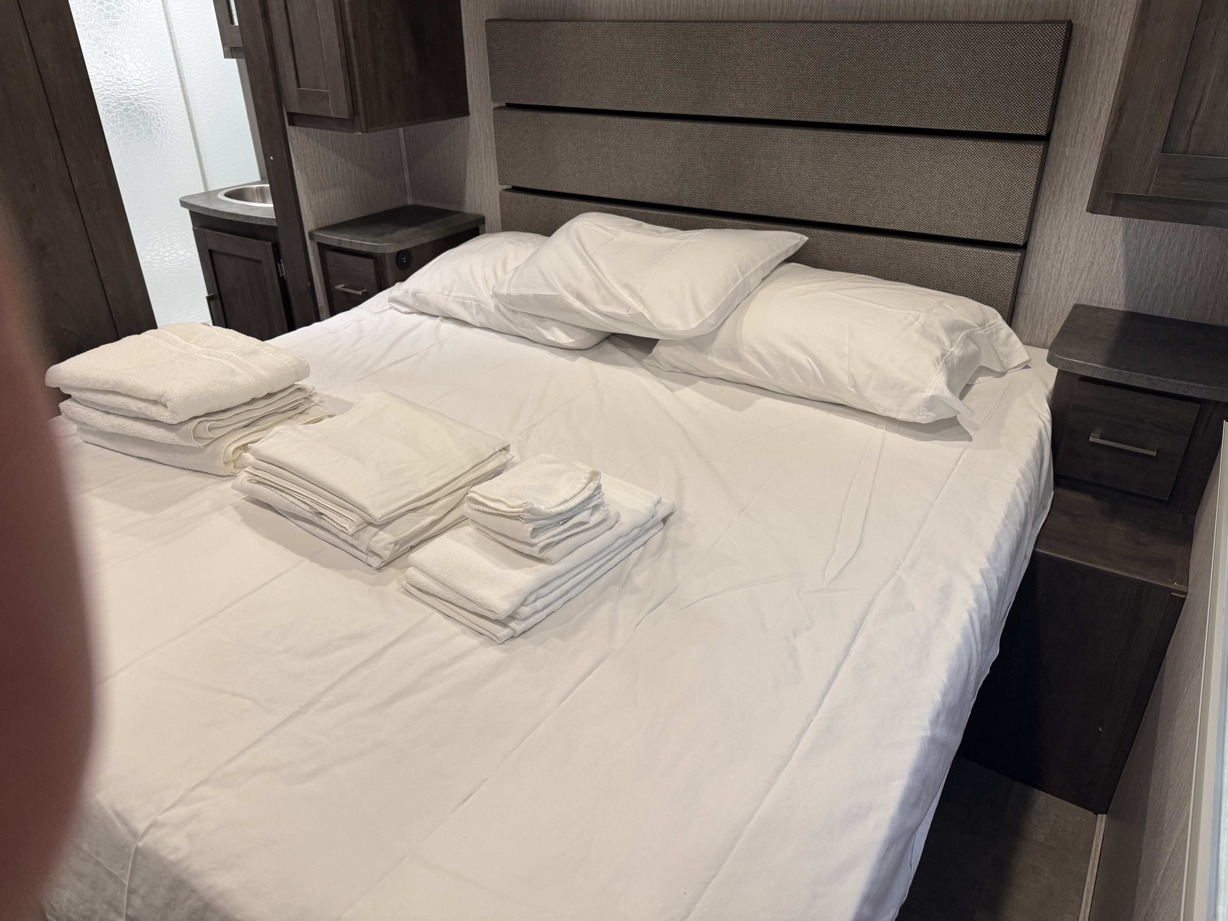 Spotless RV rental bedroom featuring a bed with white linens & stacked towels, nightstands, and an adjacent bathroom with a sink.. Coachmen Pursuit BH31 2023