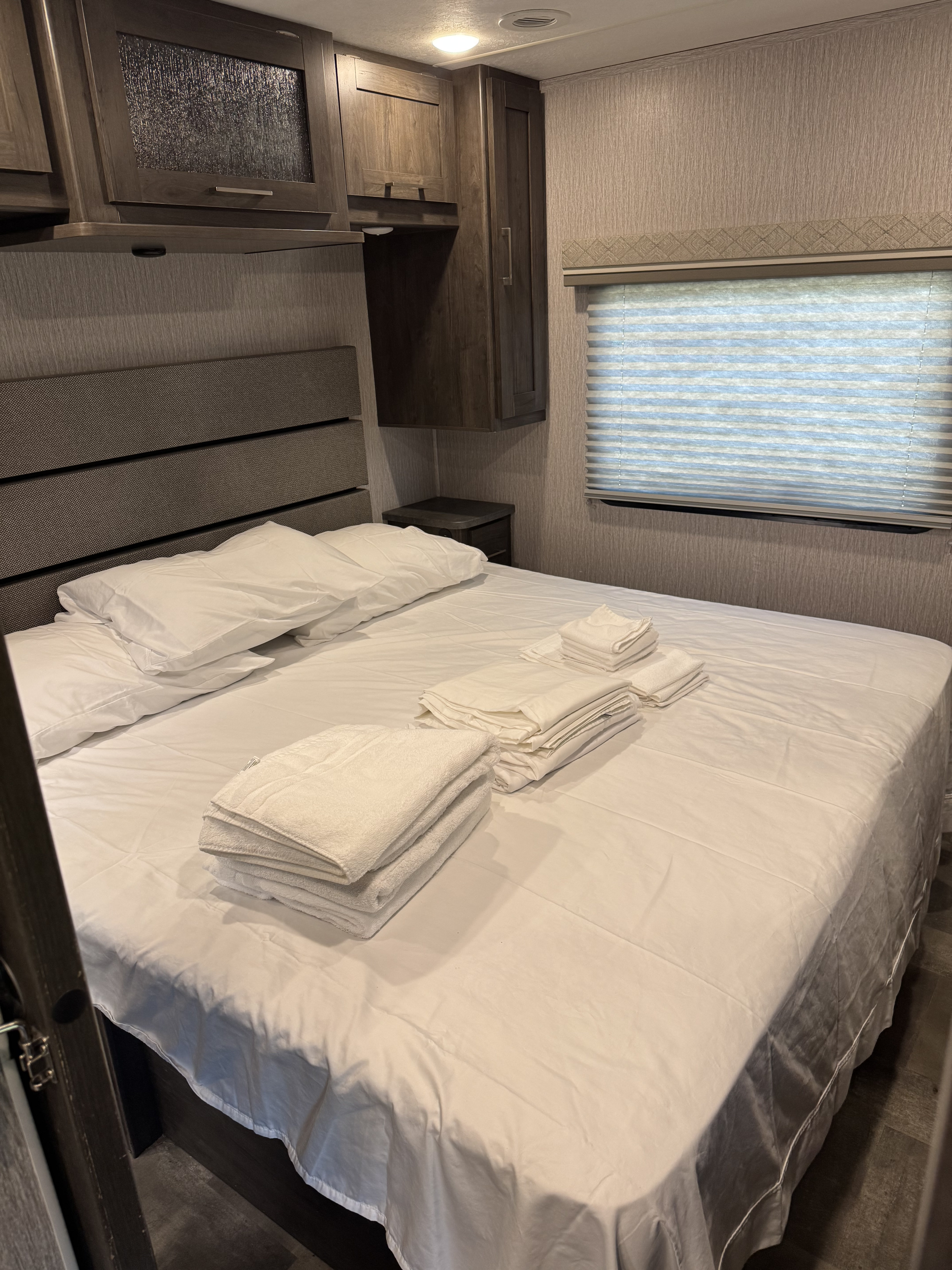 Cozy RV rental bedroom setup with fresh white bedding, neatly stacked towels, cabinets, and window blinds for comfort.. Coachmen Pursuit BH31 2023