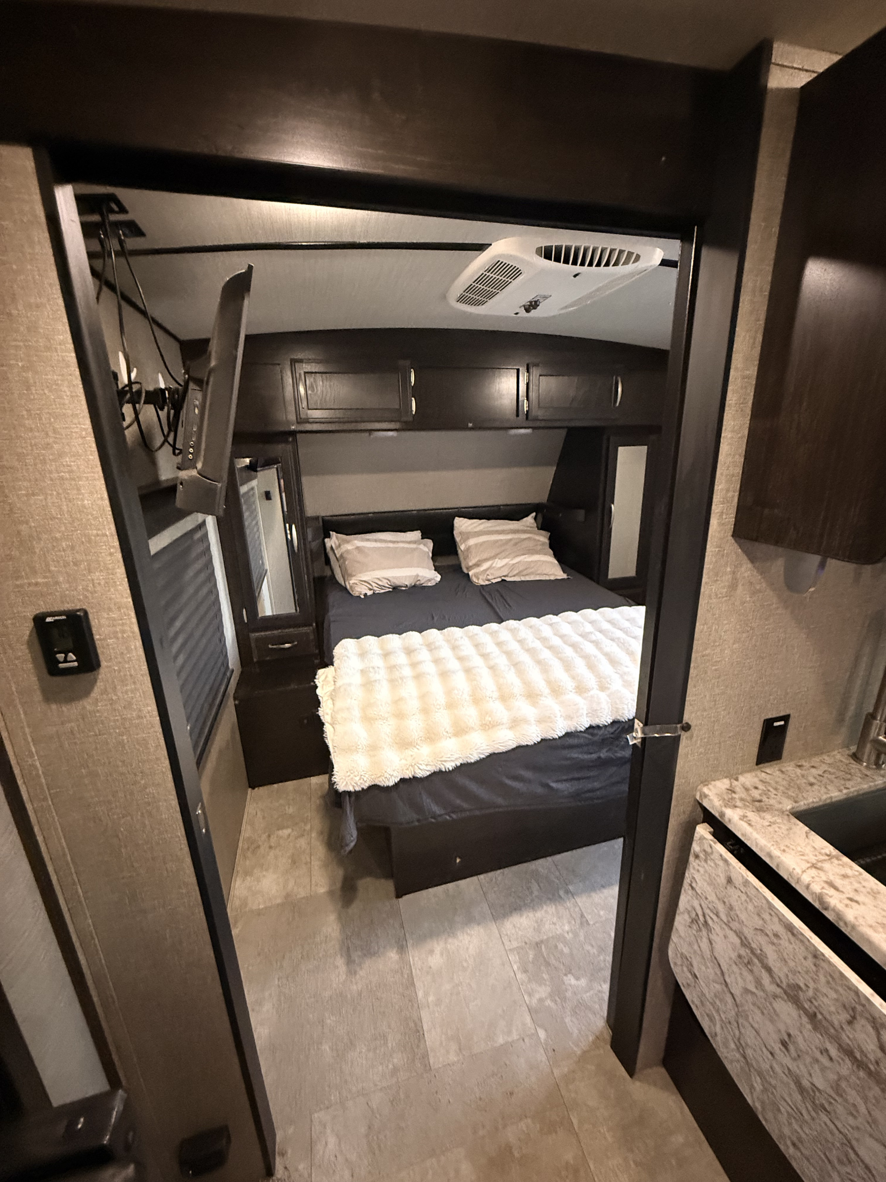 This RV rental bedroom features a bed with white blanket, overhead storage, AC, and a mounted TV, seen through a doorway.. Imagine Grand Deigns 2019