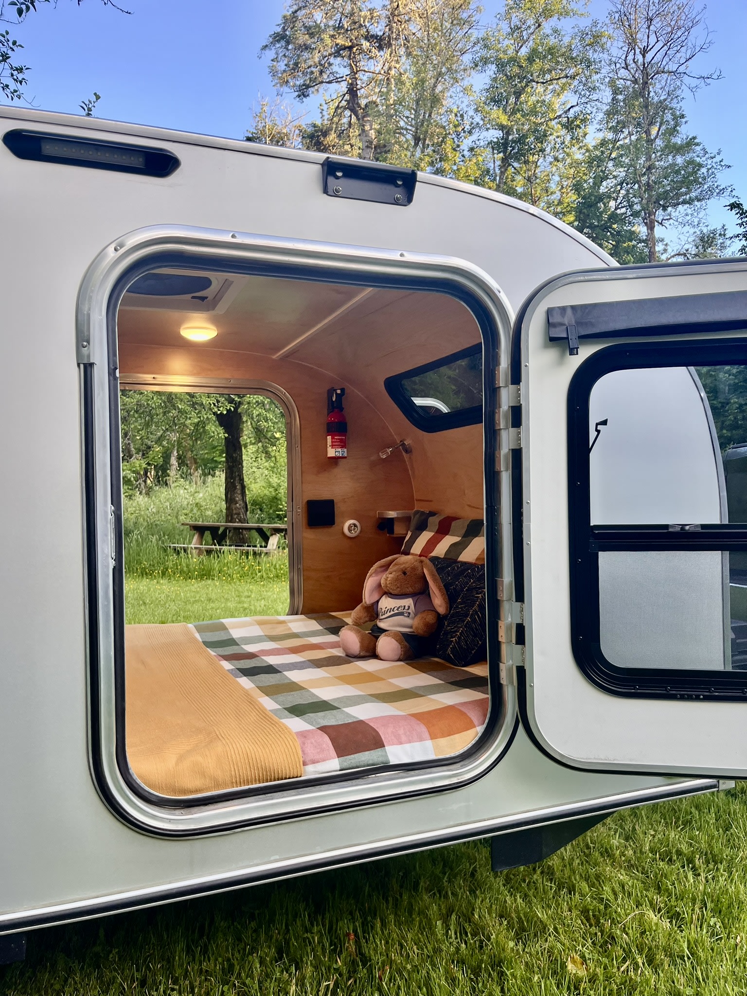 Cozy RV rental: a plush bed, bunny, and fire extinguisher inside, overlooking a green forest and picnic table.. Aero Steel 2022