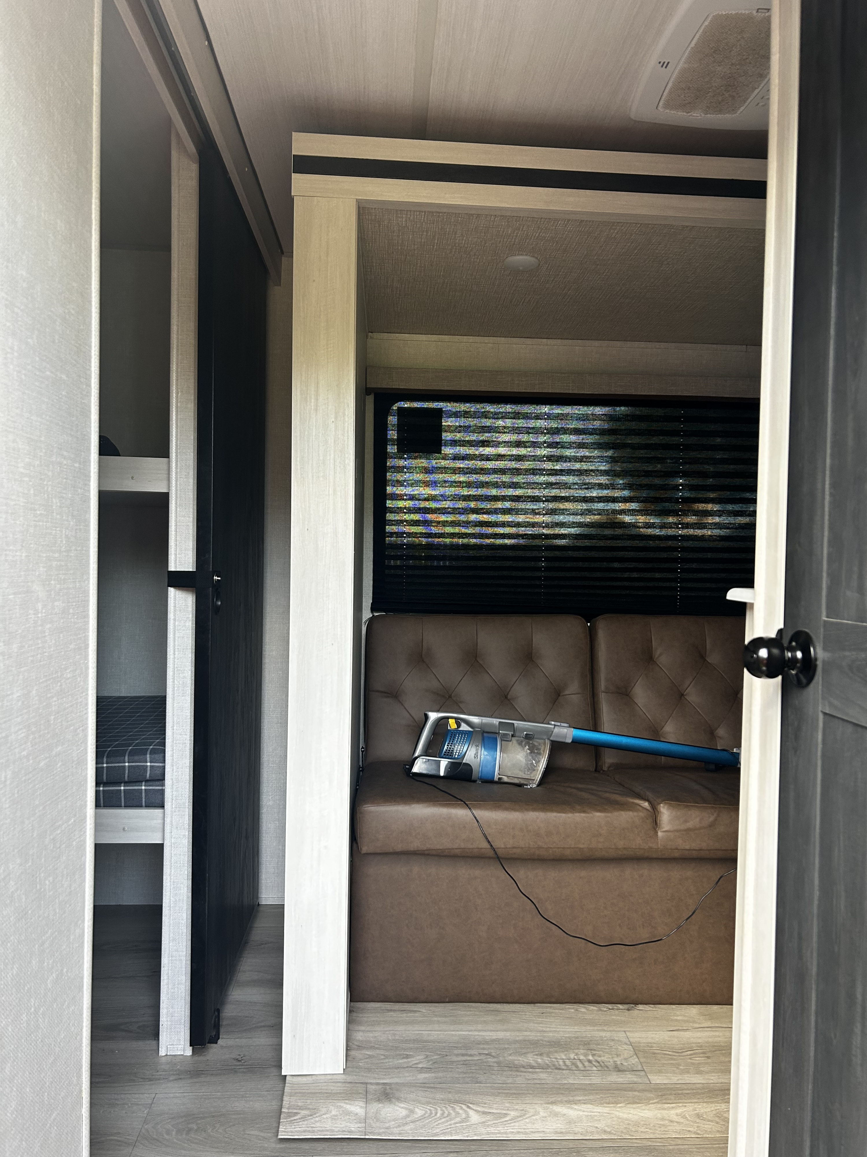RV rental interior: a tufted brown sofa with a stick vacuum, bunk beds, and a window with blinds, ready for travel.. Forest River Surveyor Legend X 2025