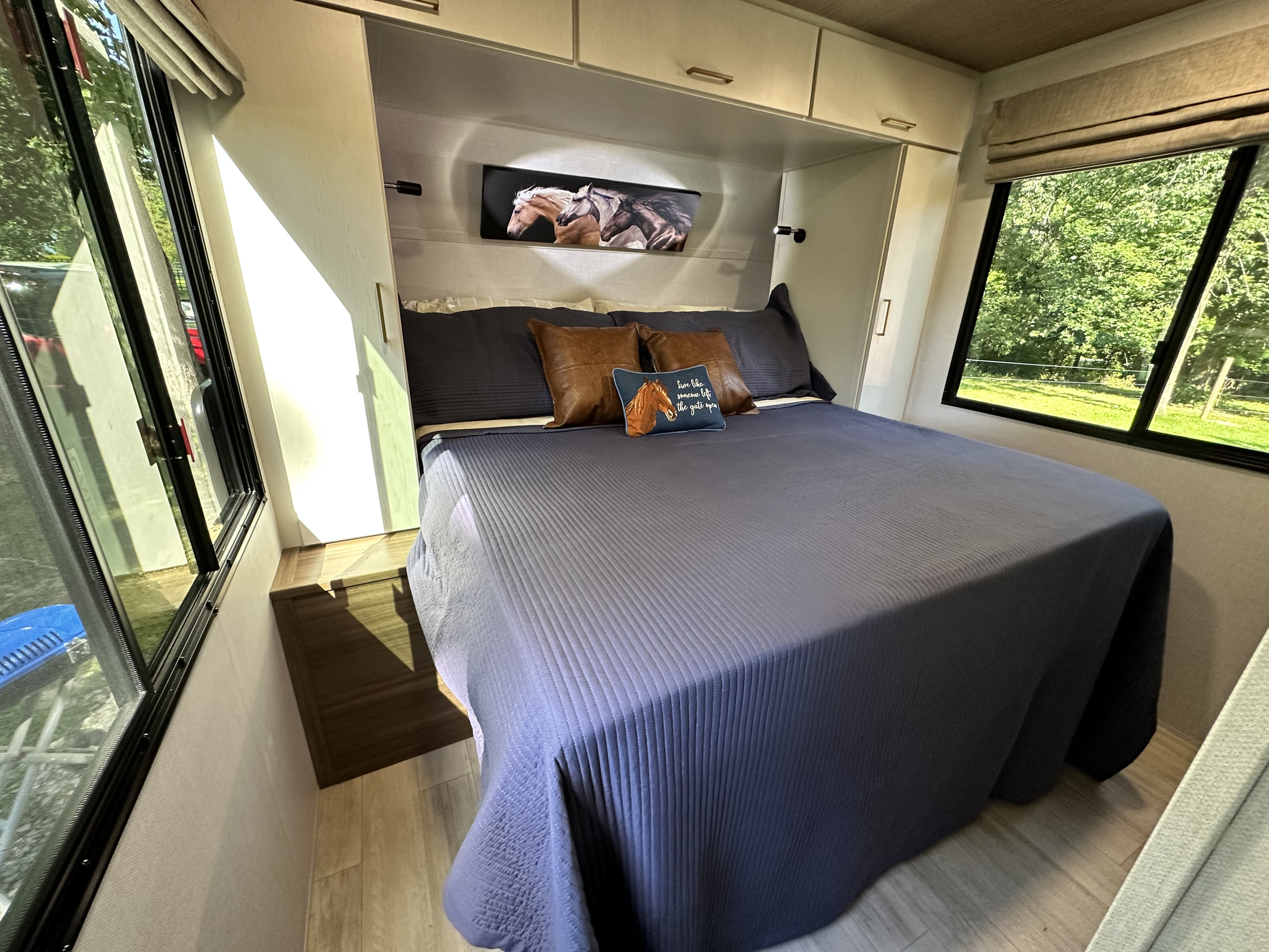 Cozy RV rental bedroom featuring blue bedding, horse art, overhead storage, and sunlit windows with nature views.. Forest River Vibe 2026