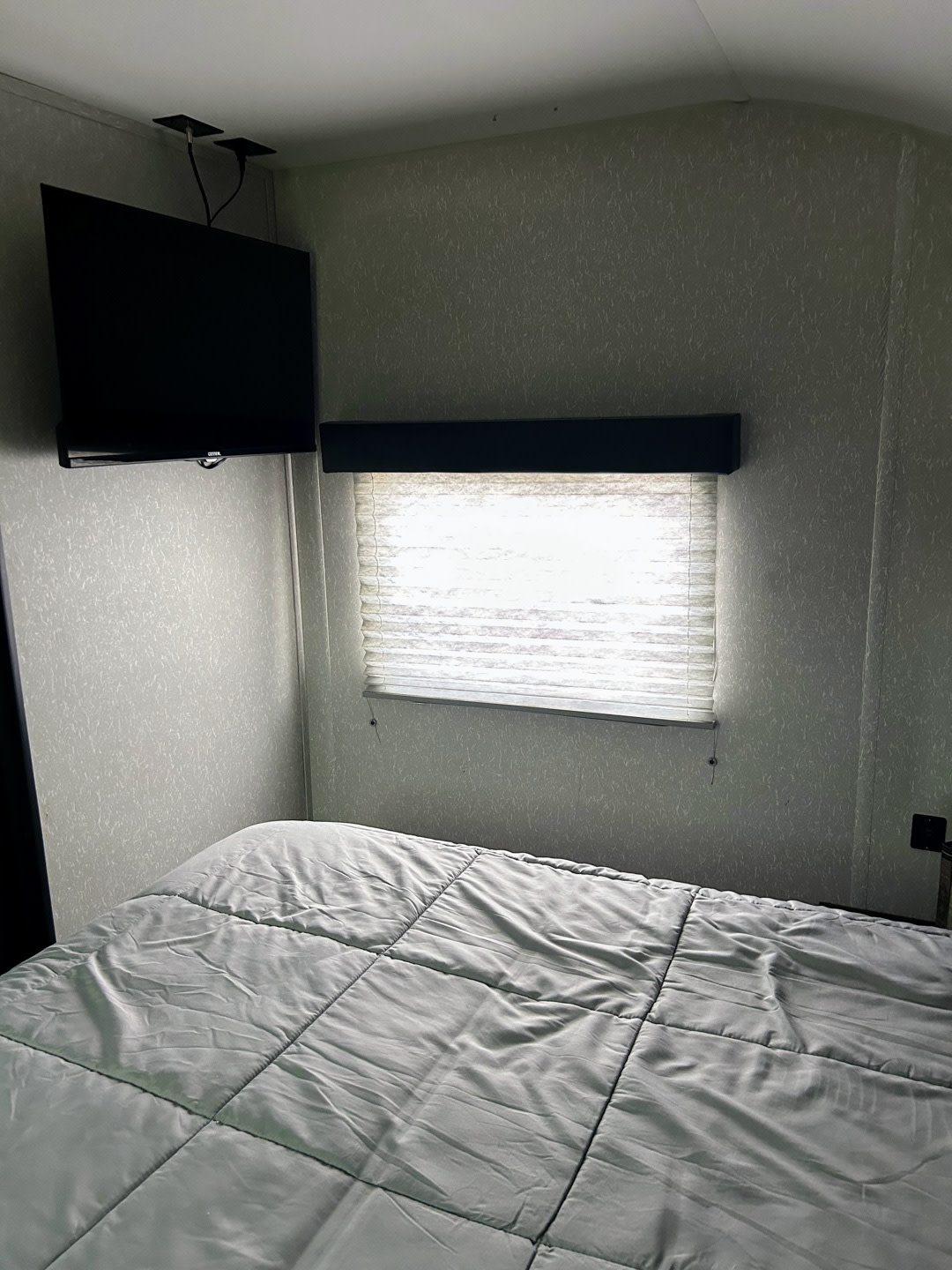 This RV rental bedroom features a cozy bed with light gray comforter, a window with blinds, and a wall-mounted TV, ideal for compact living.. Forest River Cherokee Grey Wolf 2022