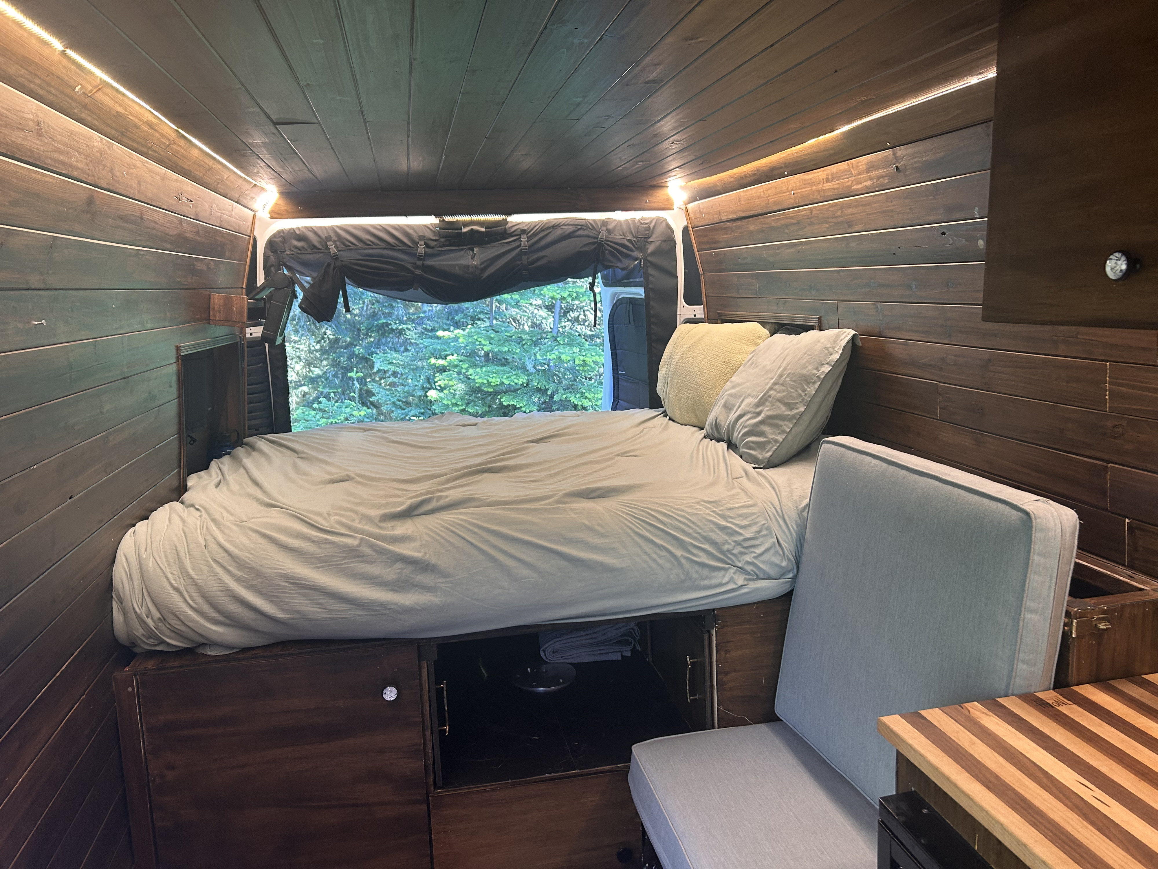 Cozy RV rental interior with wood-paneled walls, bed, ambient strip lighting, window view of trees, cushioned seat, & storage.. RAM Promaster 2022