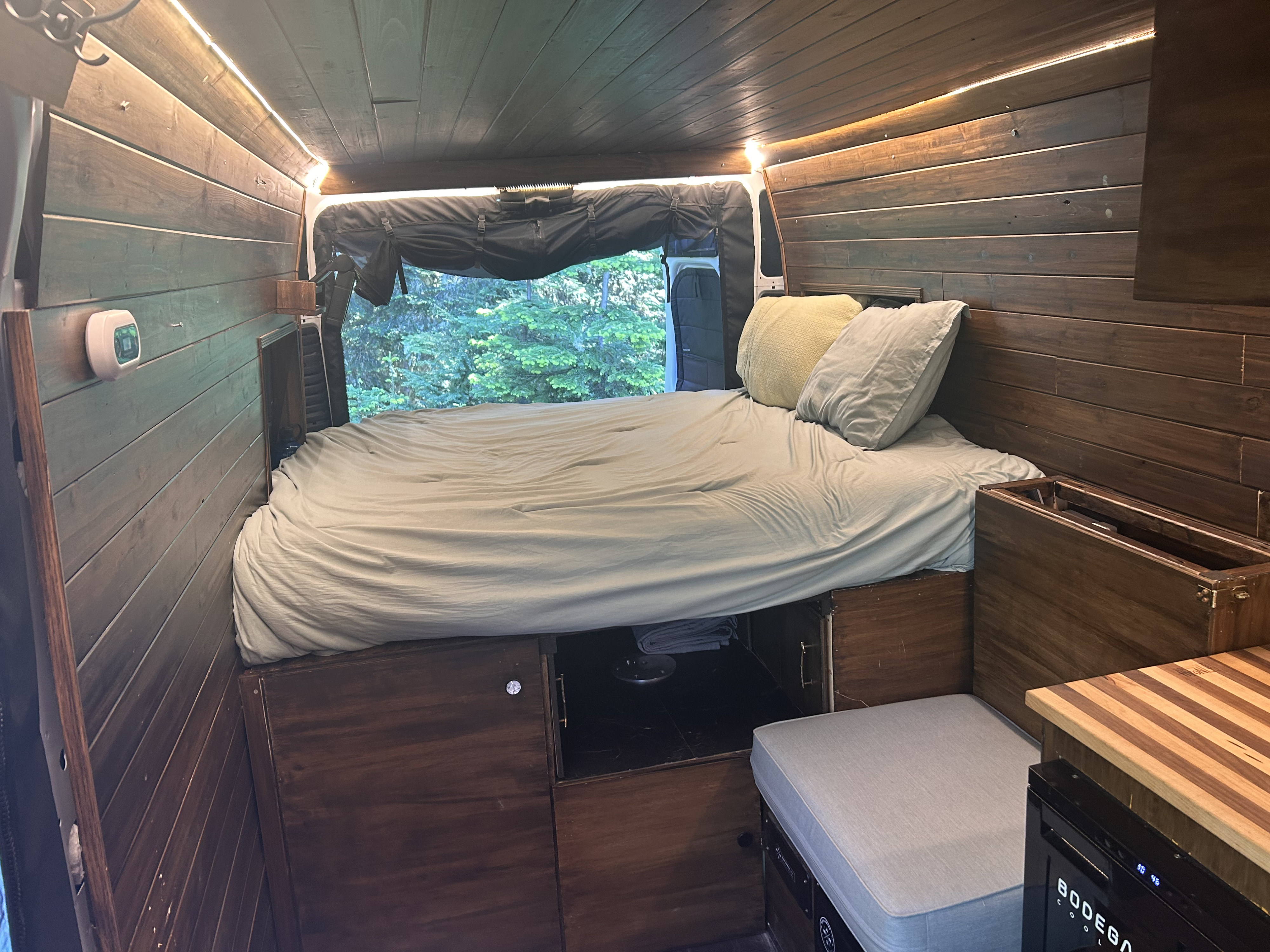 This RV rental's cozy interior features a comfortable bed, warm wooden paneling, ambient lighting, and a window showing lush trees.. RAM Promaster 2022