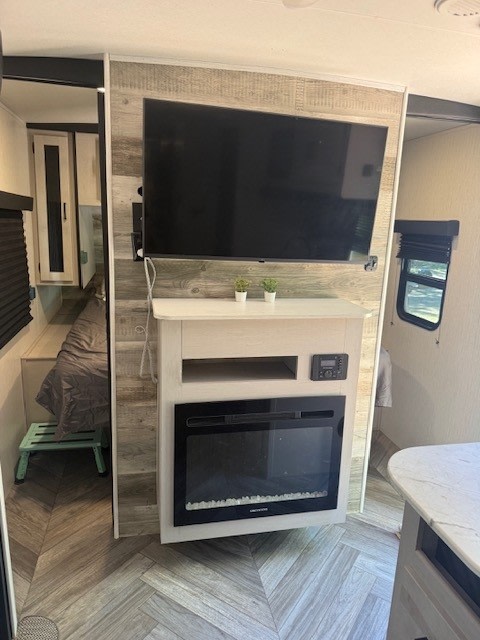 Modern RV rental living space featuring a wall-mounted TV, electric fireplace, and herringbone floors. Bedroom left.. Heartland RVs Mallard 2023