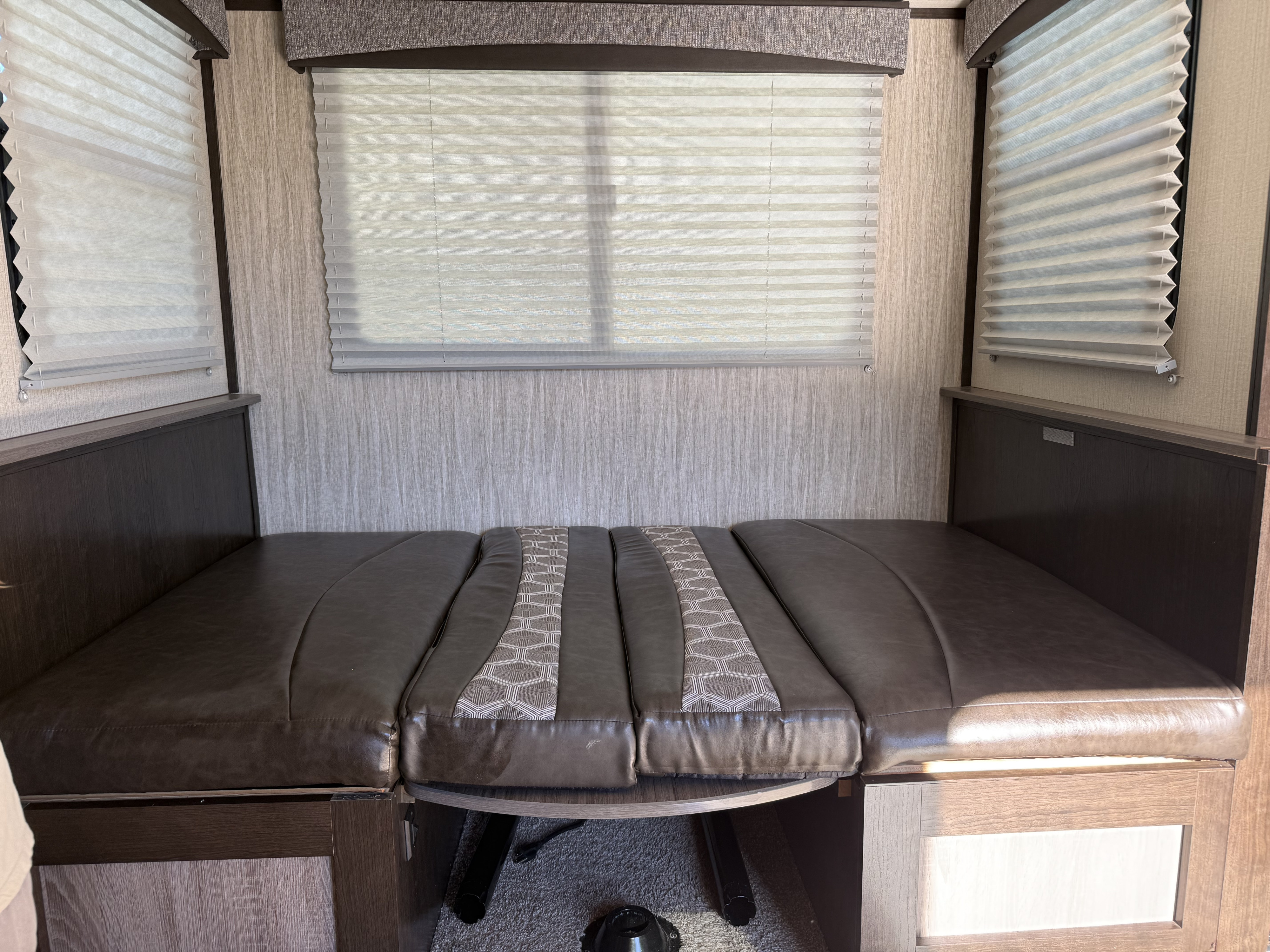 RV rental dinette converts to a bed, featuring dark cushions, light wood walls, and pleated window blinds.. Cruiser RV Shadow Cruiser 2018