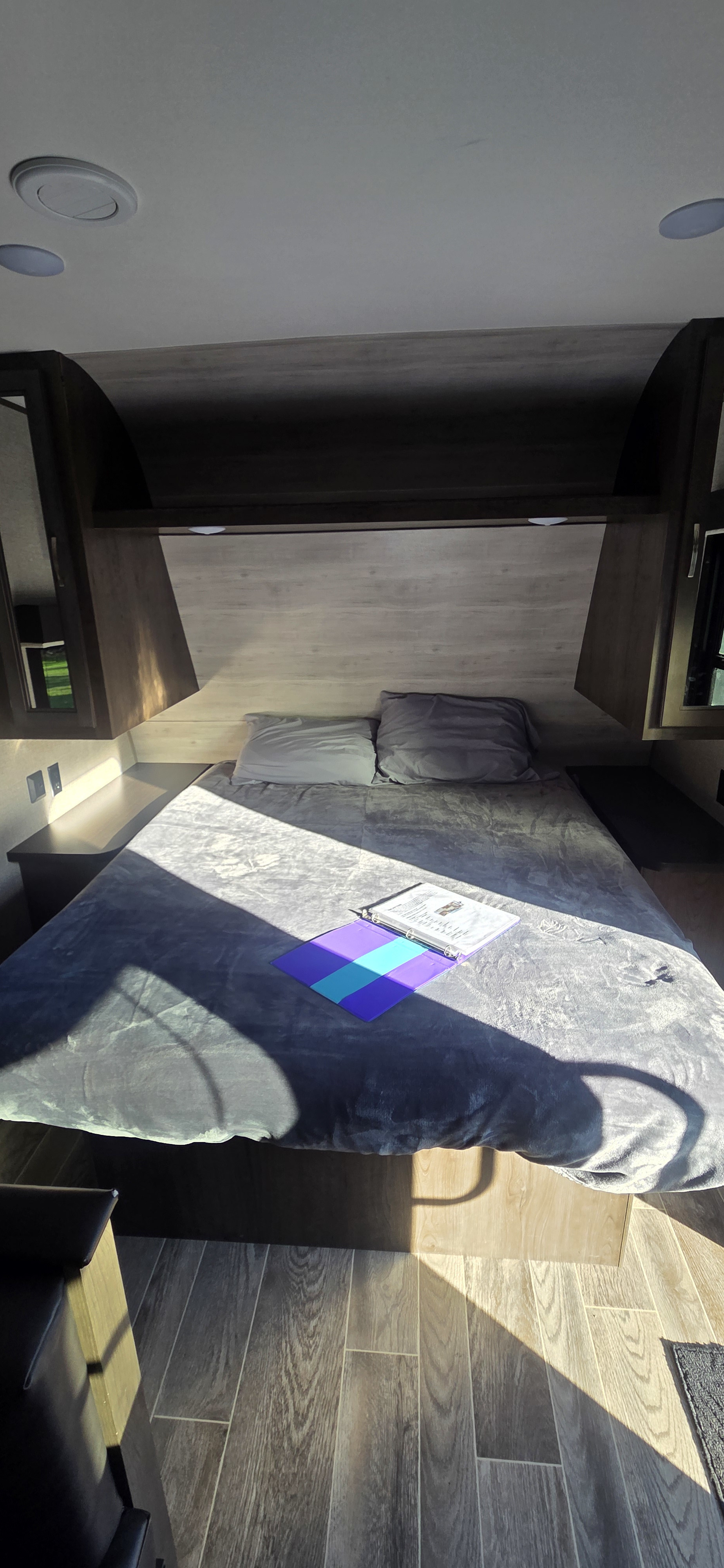 Interior of an RV rental bedroom with a grey bed, overhead cabinets, and sunlit wood-look floor.. Jayco Jay Flight SLX 2021