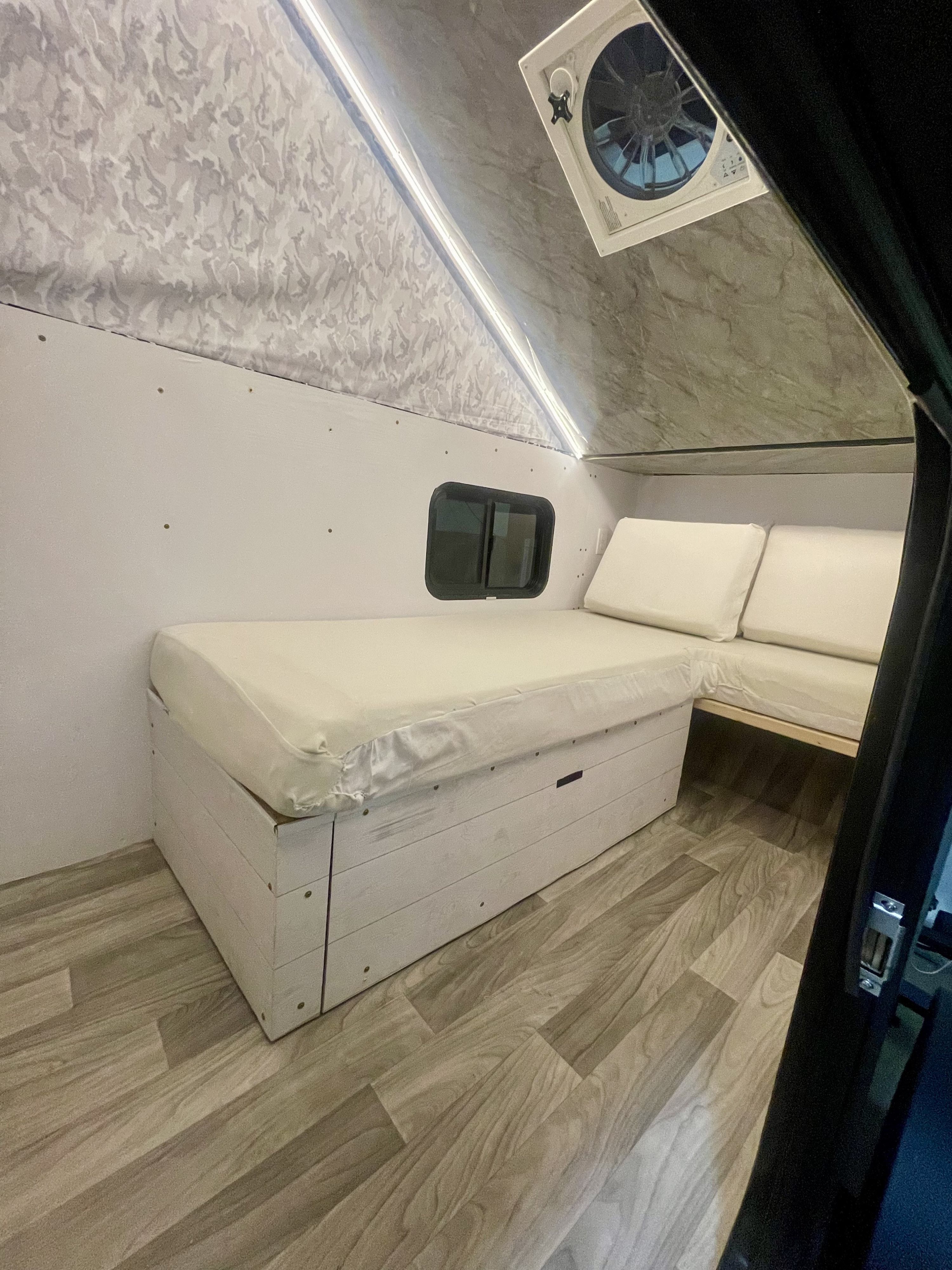 Cozy RV rental interior featuring a white mattress bed with storage, L-shaped seating, small window, and wood-look floor.. Homemade Box 2020
