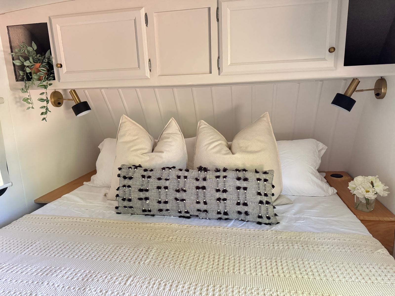 Cozy RV rental bedroom with white bedding, cream pillows, a textured grey pillow, brass wall lights, and plant details.. Coachmen Apex 2017