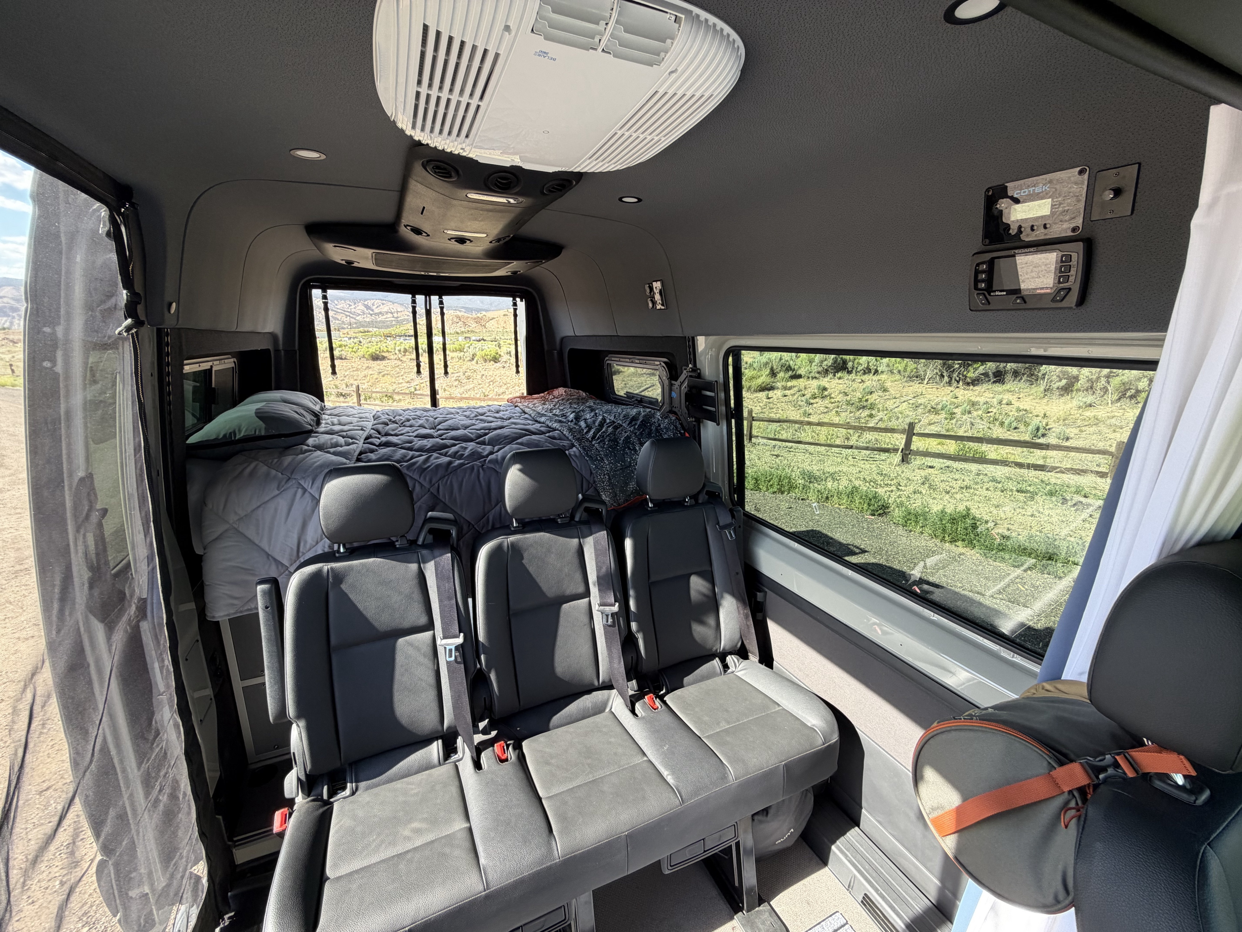 Inside this rv rental: three seats, a cozy bed, and nature views from panoramic windows. Ceiling AC and control panels visible.. Mercedes-Benz Sprinter 2022