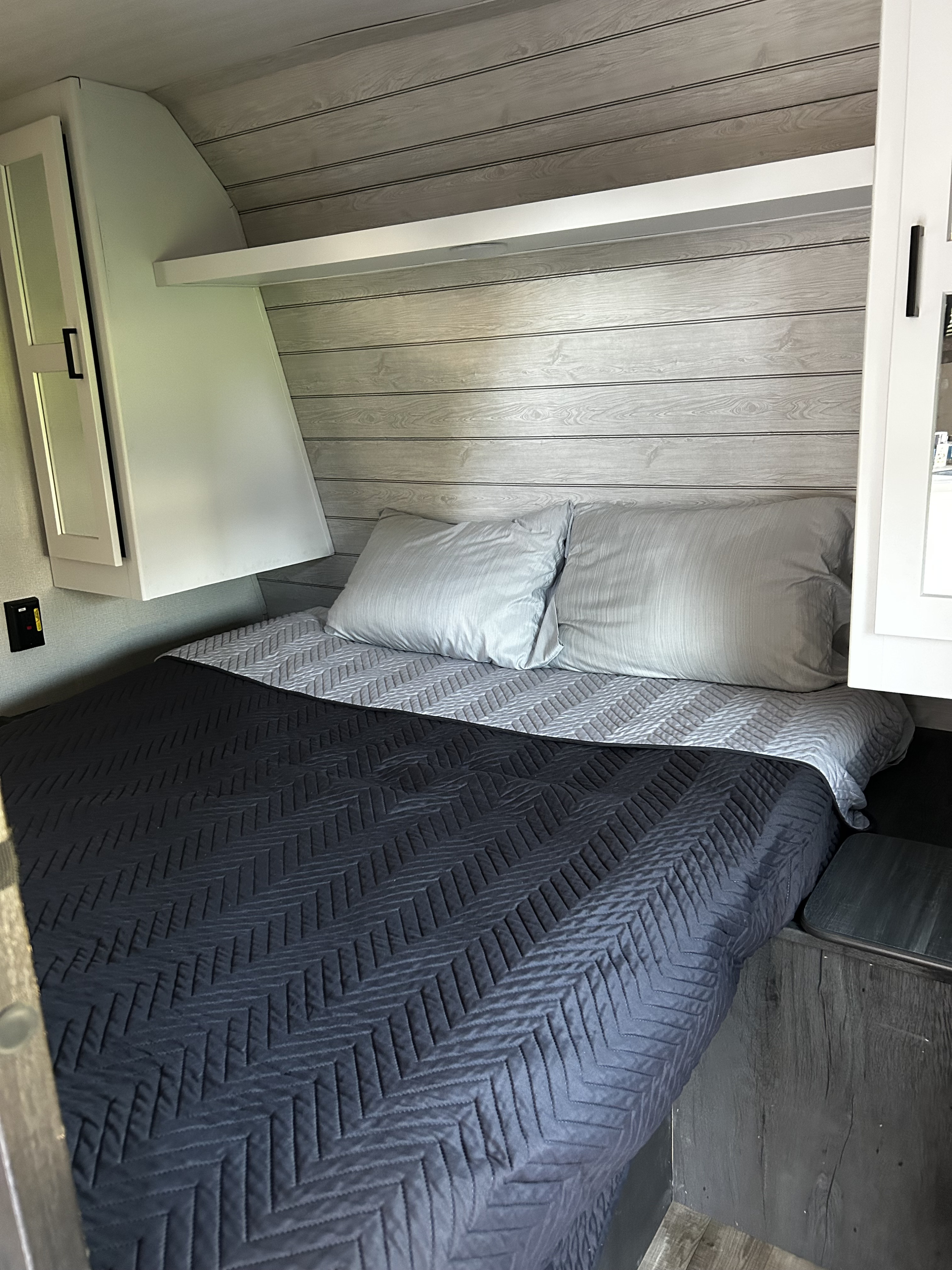 RV rental bedroom featuring a cozy bed with dark blue quilt, pillows, and wood-paneled walls with shelving.. Keystone RV Hideout 2023