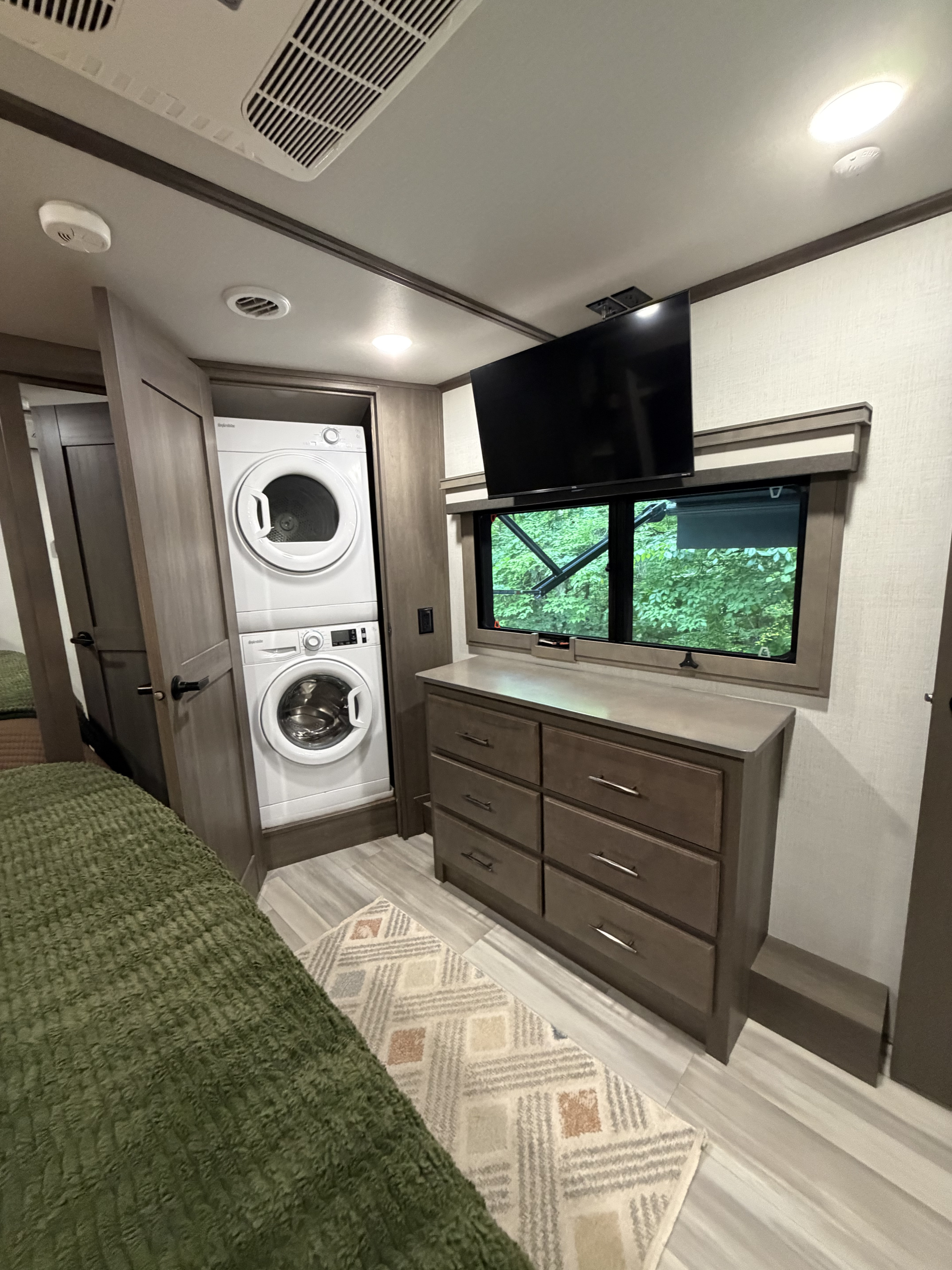 RV rental bedroom with stacked washer/dryer, dresser, TV, and a window overlooking green trees. Features a green bedspread and rug.. Grand Design Solitude 2024