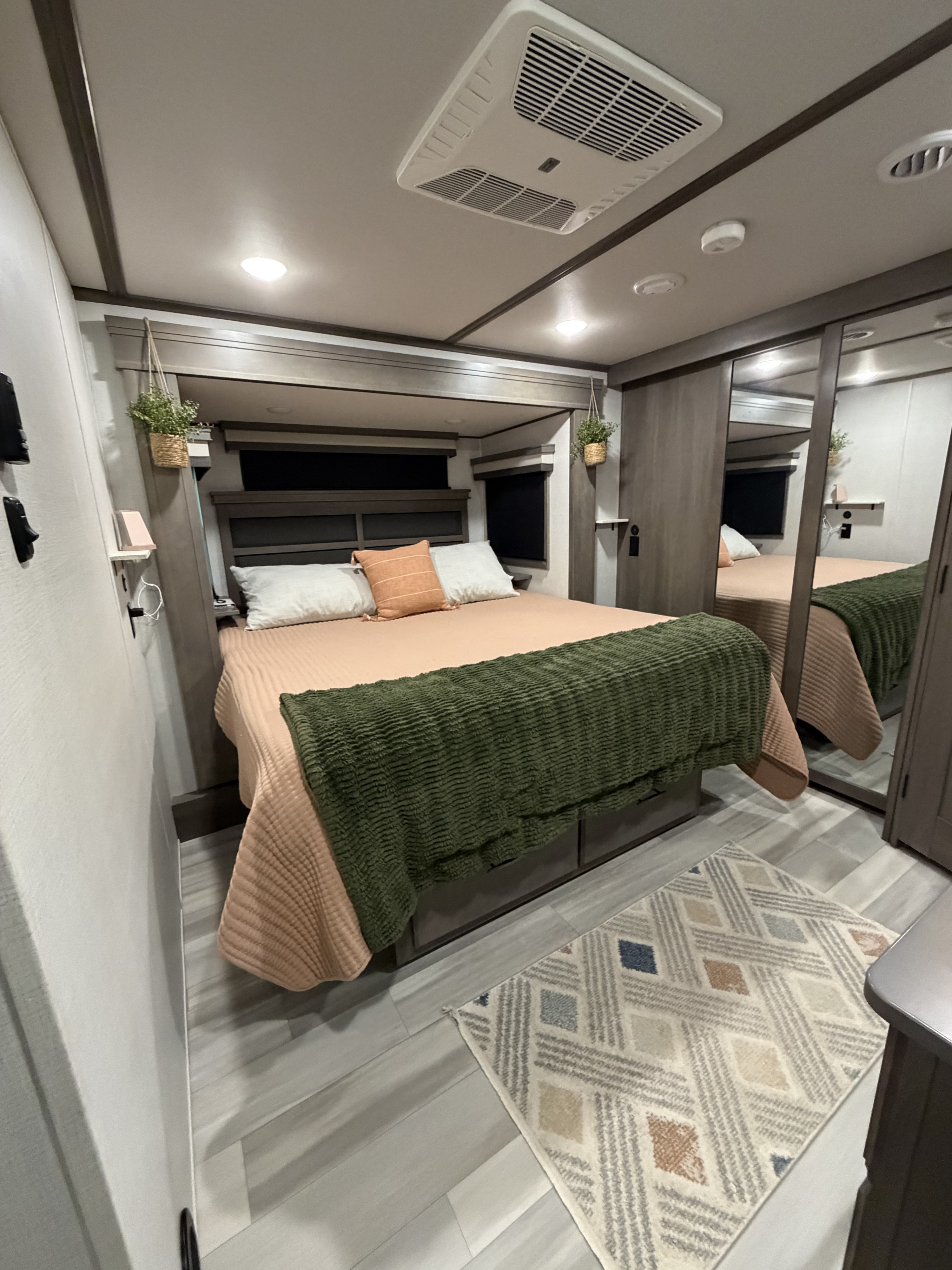 Modern RV rental bedroom: cozy bed with peach and green linens, mirrored closet, patterned rug, and ceiling AC.. Grand Design Solitude 2024