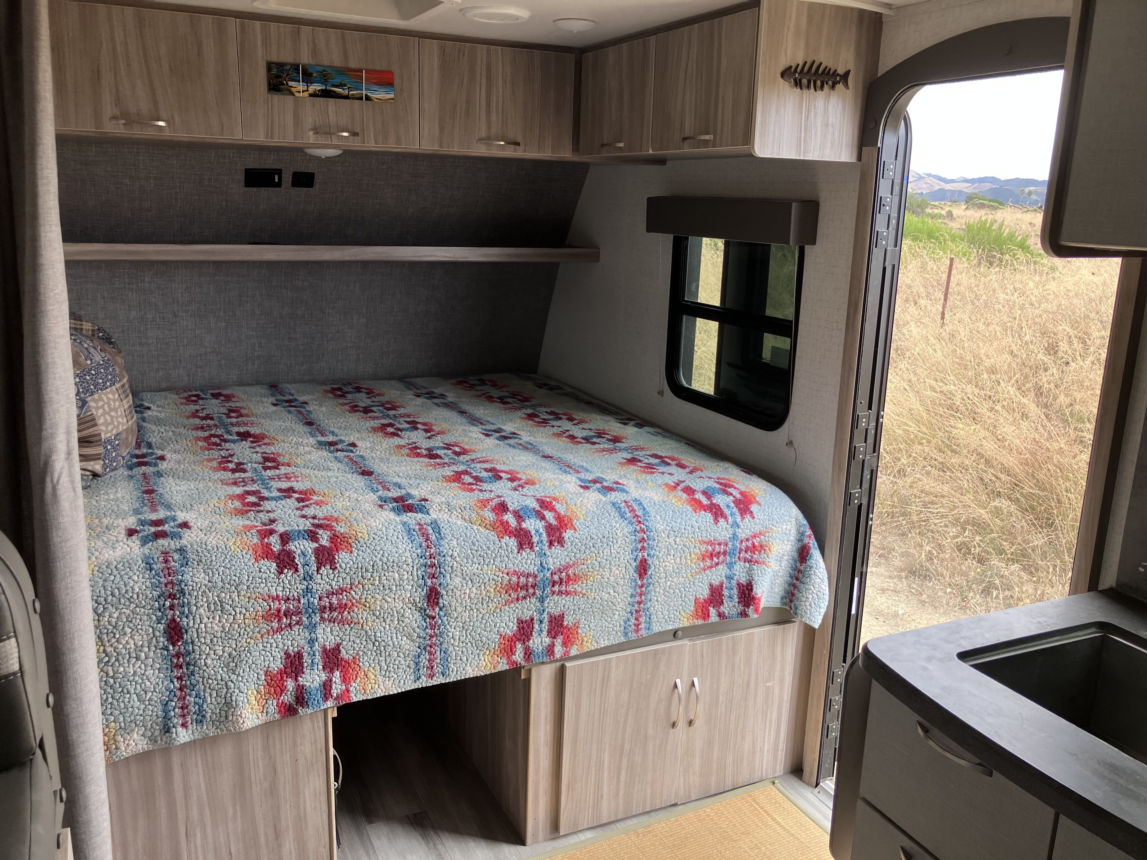 Cozy RV rental bedroom with colorful bed, storage, cabinets, and open door view of hills and dry grass.. Winnebago Micro Winnie 2021