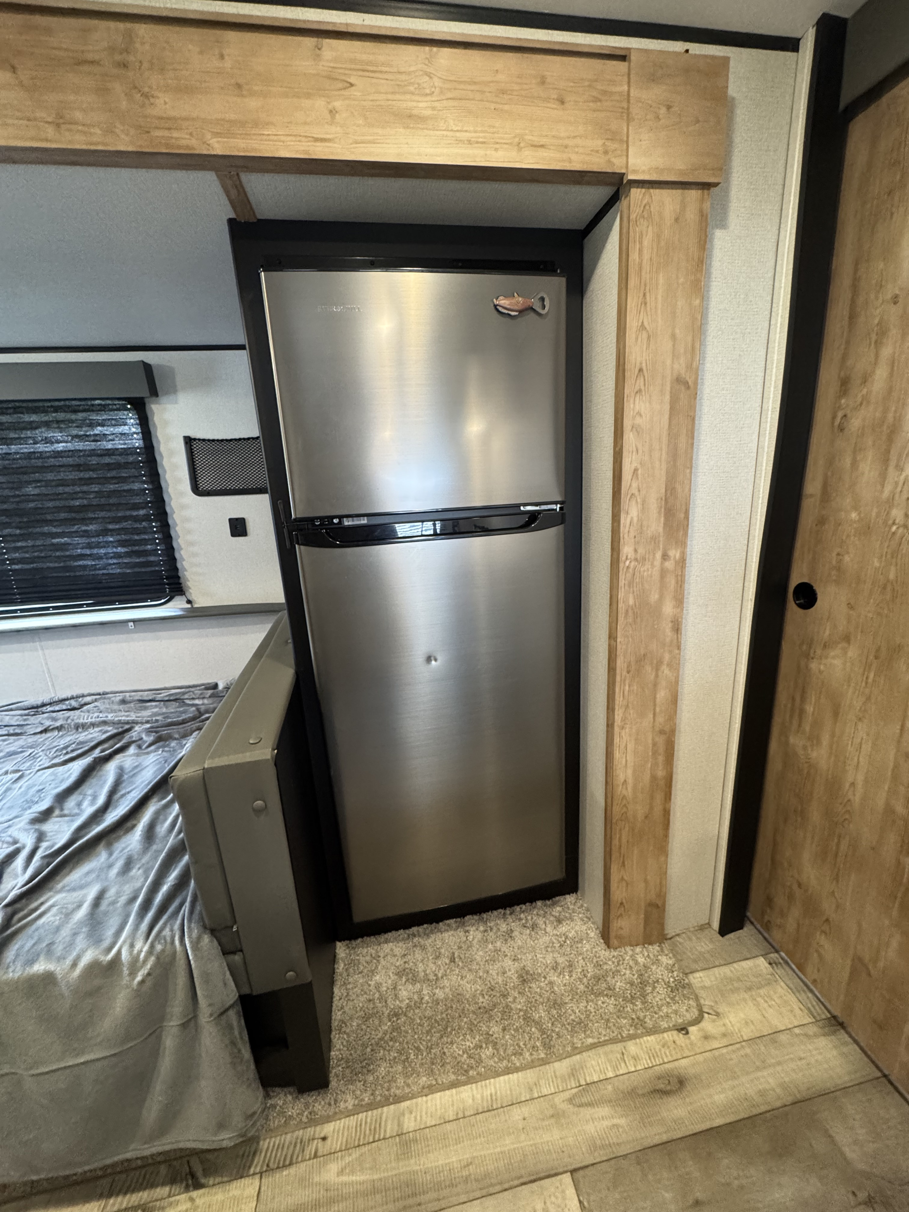 Inside an RV rental: a stainless steel refrigerator next to a cozy sleeping area, with wood accents and window blinds.. Keystone RV Springdale 2022