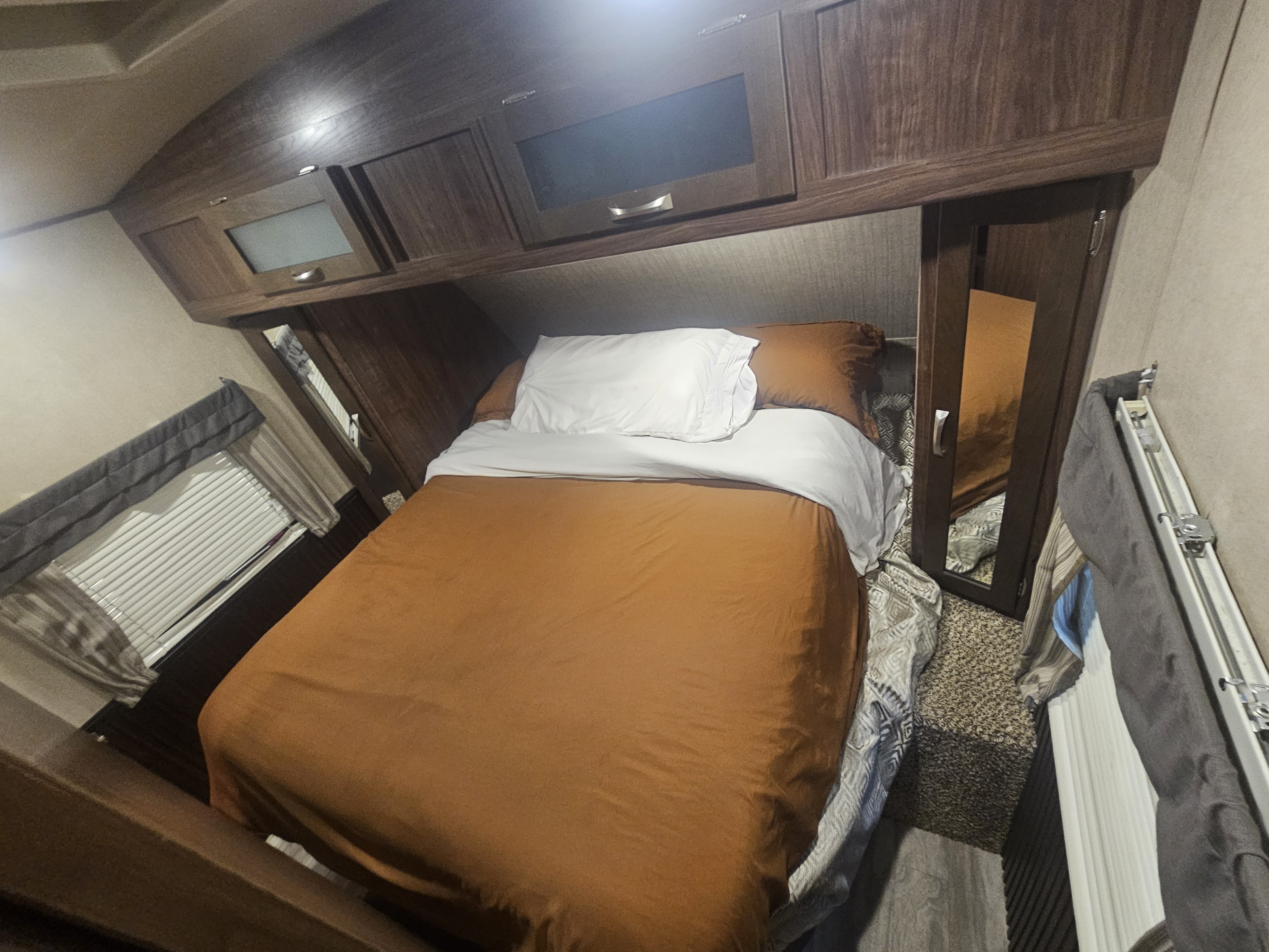 Cozy rv rental bedroom with an inviting orange comforter, white pillows, overhead cabinets, and a window with blinds.. Dutchmen 242bhsl 2017