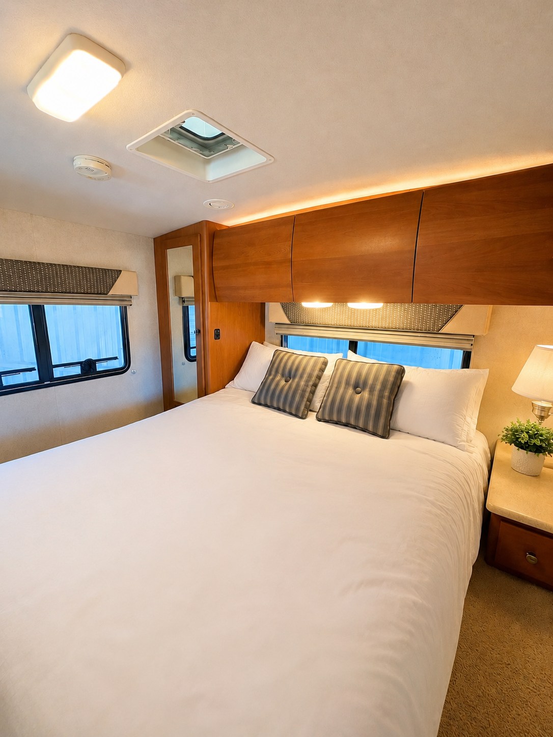 This RV rental bedroom features a comfy queen bed, overhead wooden cabinets, bright ceiling lights, a window, and nightstand.. Winnebago Outlook 2008