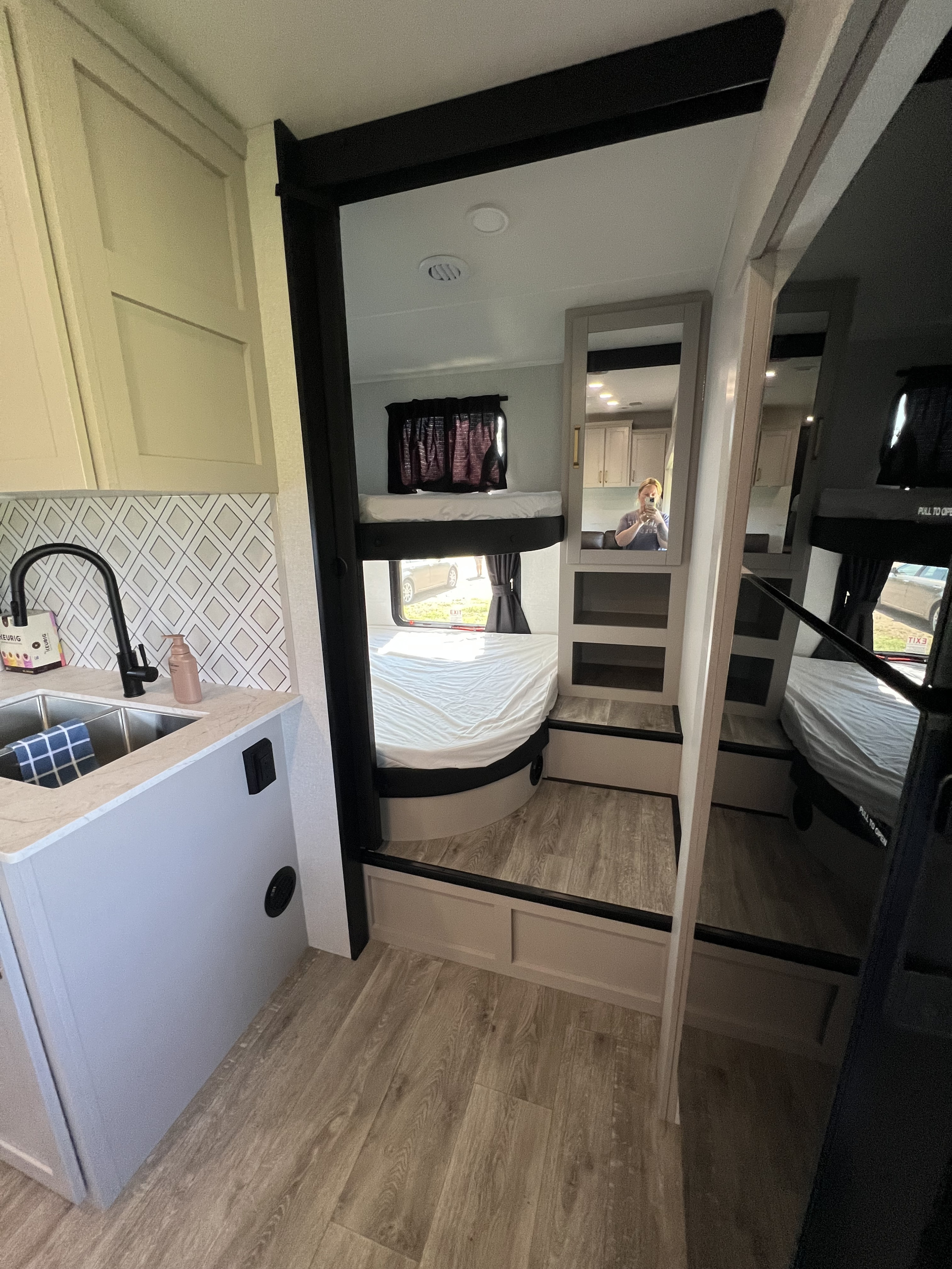 Bright RV rental interior: modern kitchen with sink on left, mirror reflecting a person, and cozy bunk beds on the right. Light wood flooring.. Sundance xlt 28DB 2026