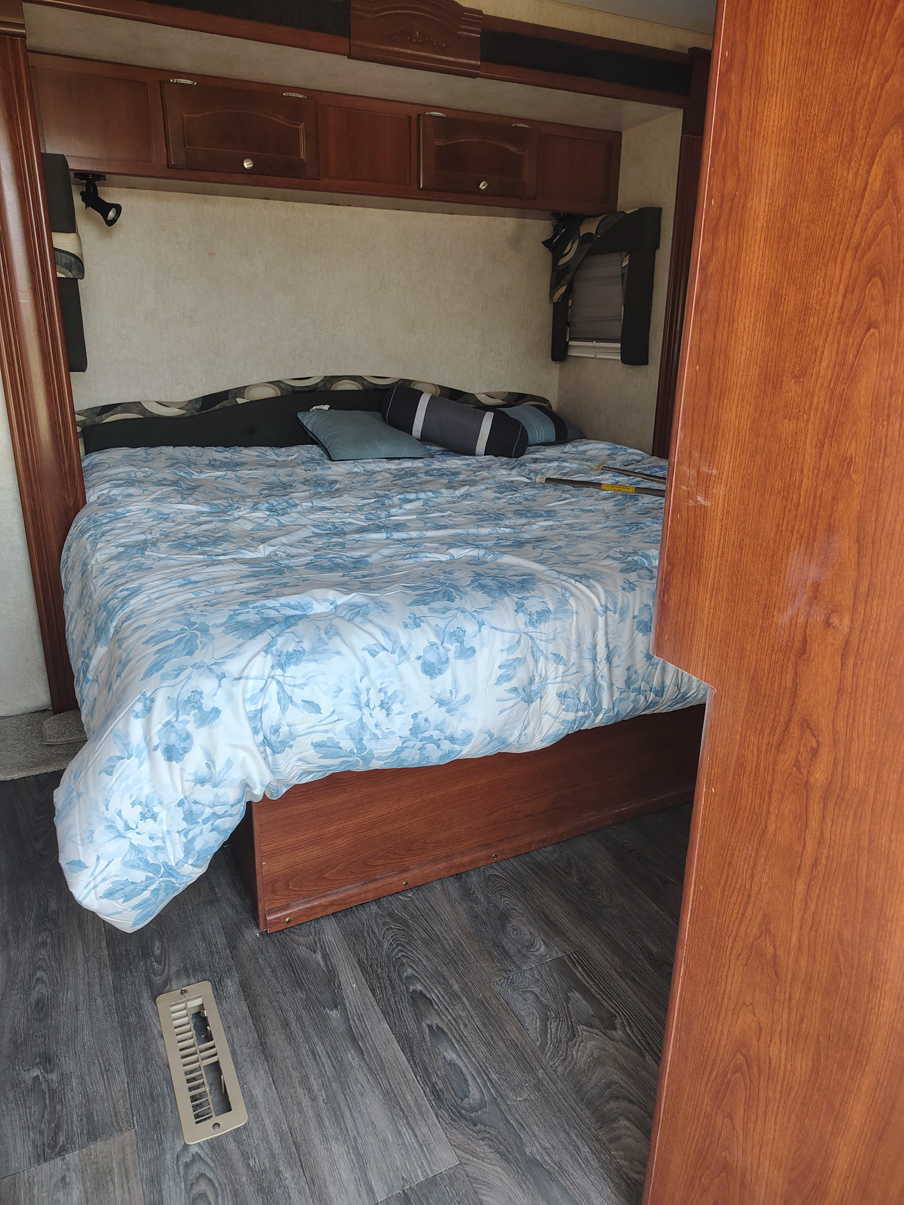 Compact RV rental bedroom featuring a bed with a blue floral duvet, overhead wood cabinets, and dark laminate flooring.. Fleetwood Terry 2007