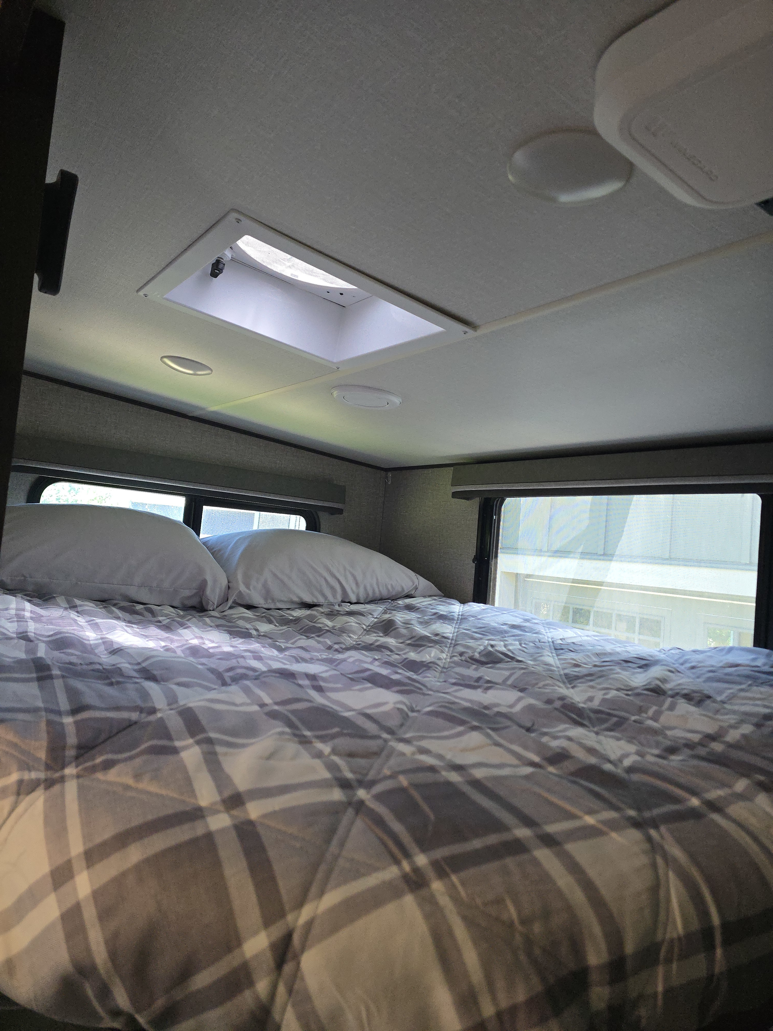 Cozy RV rental bedroom featuring a comfy plaid bed with pillows, large windows, and a ceiling skylight/vent overhead.. Grand Design Reflection 2021