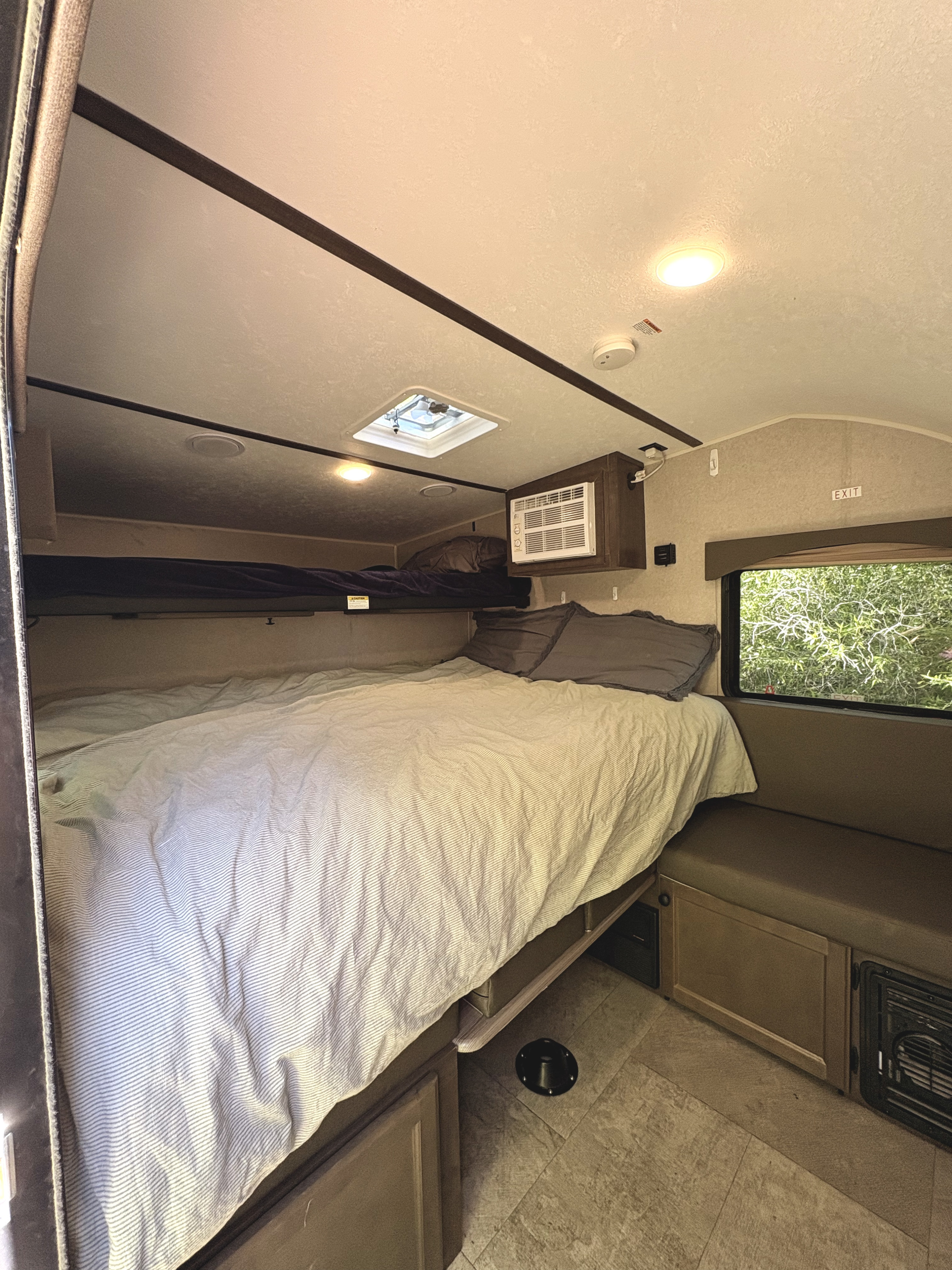 Cozy RV rental interior featuring a main bed with striped bedding, an upper bunk, and a side dinette by a window.. Sunset Park RV SunRay 2022