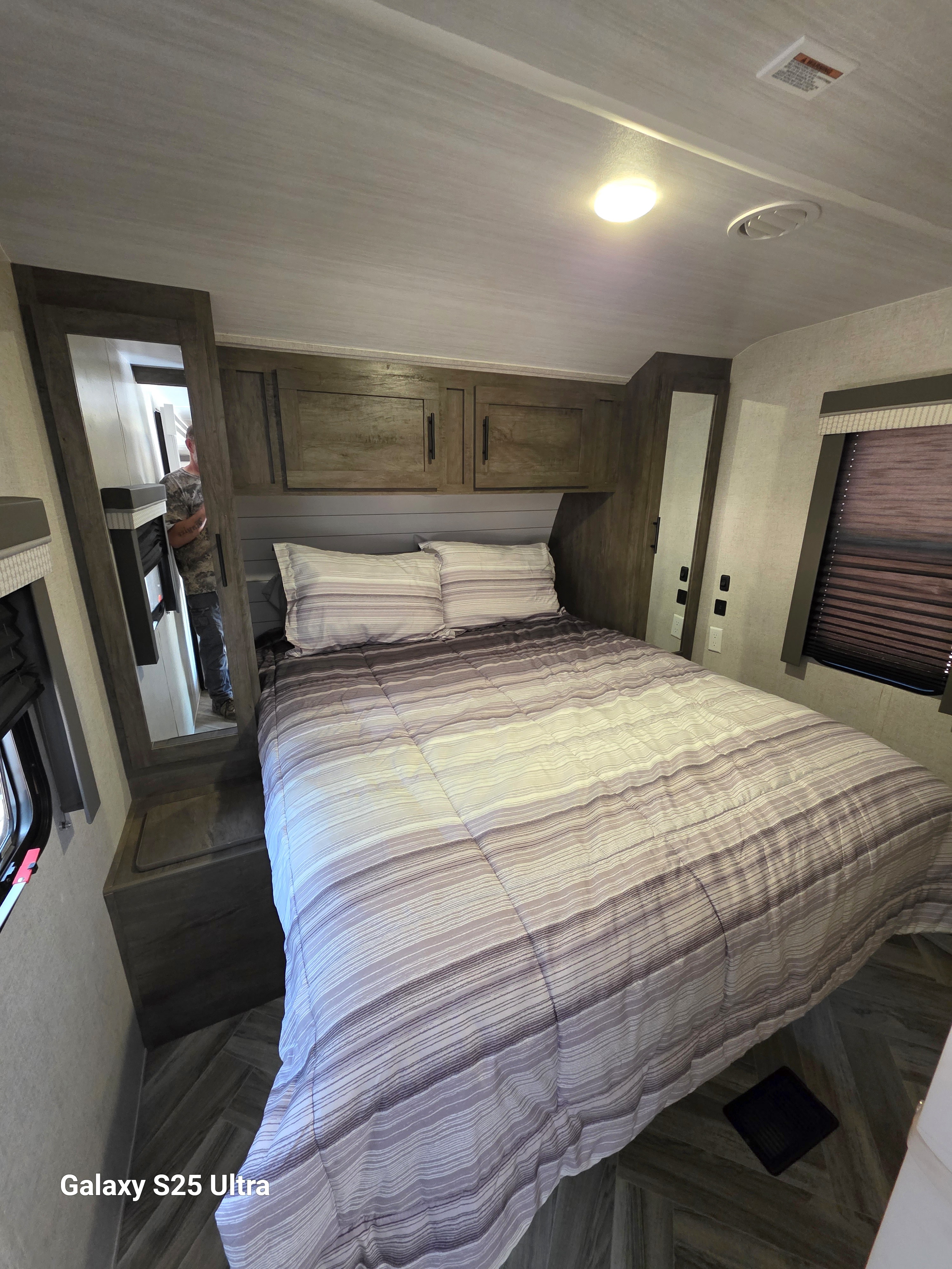 Cozy RV rental bedroom: striped bed, wood cabinets, ceiling light & vent, window with blinds. Person in mirror.. Forest River Wildwood 2021