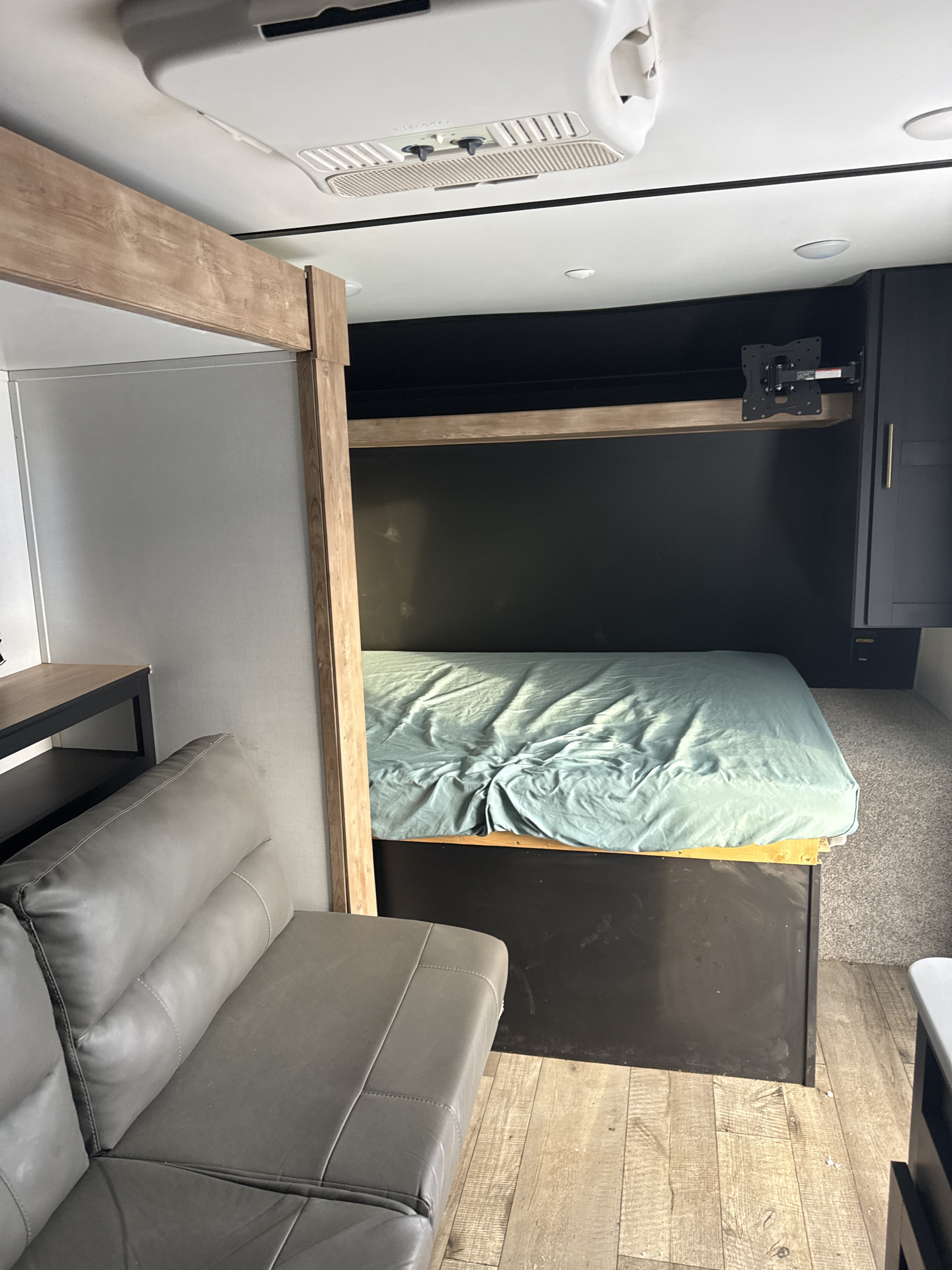 Interior of an RV rental showing a bed, gray sofa, ceiling AC unit, and a wall-mounted TV bracket.. Keystone RV Springdale 2022