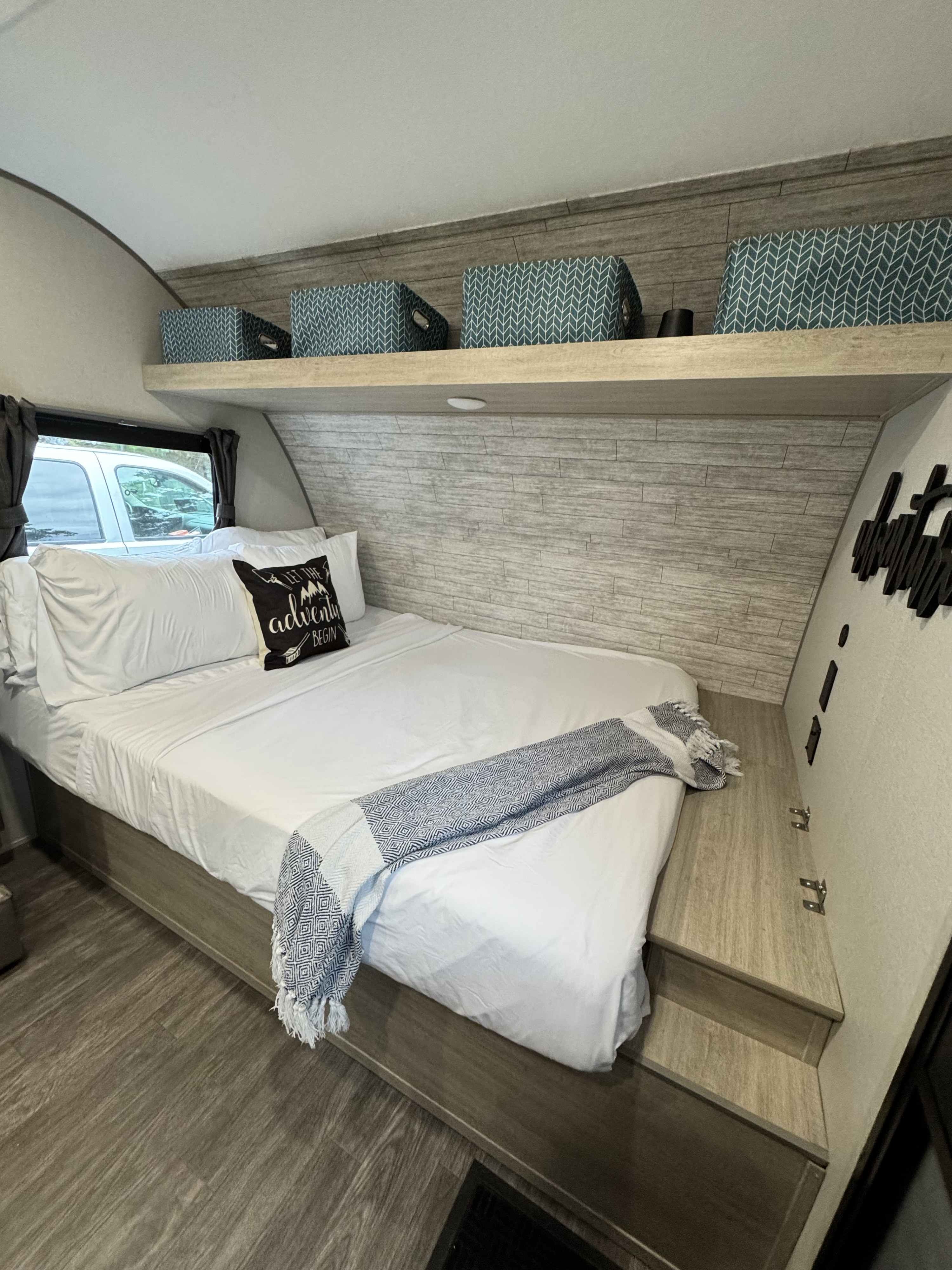 Cozy RV rental bedroom with white bedding, an "adventure" pillow, patterned throw, shelf storage, and wood accents.. Forest River Aurora Aurora Light 2026