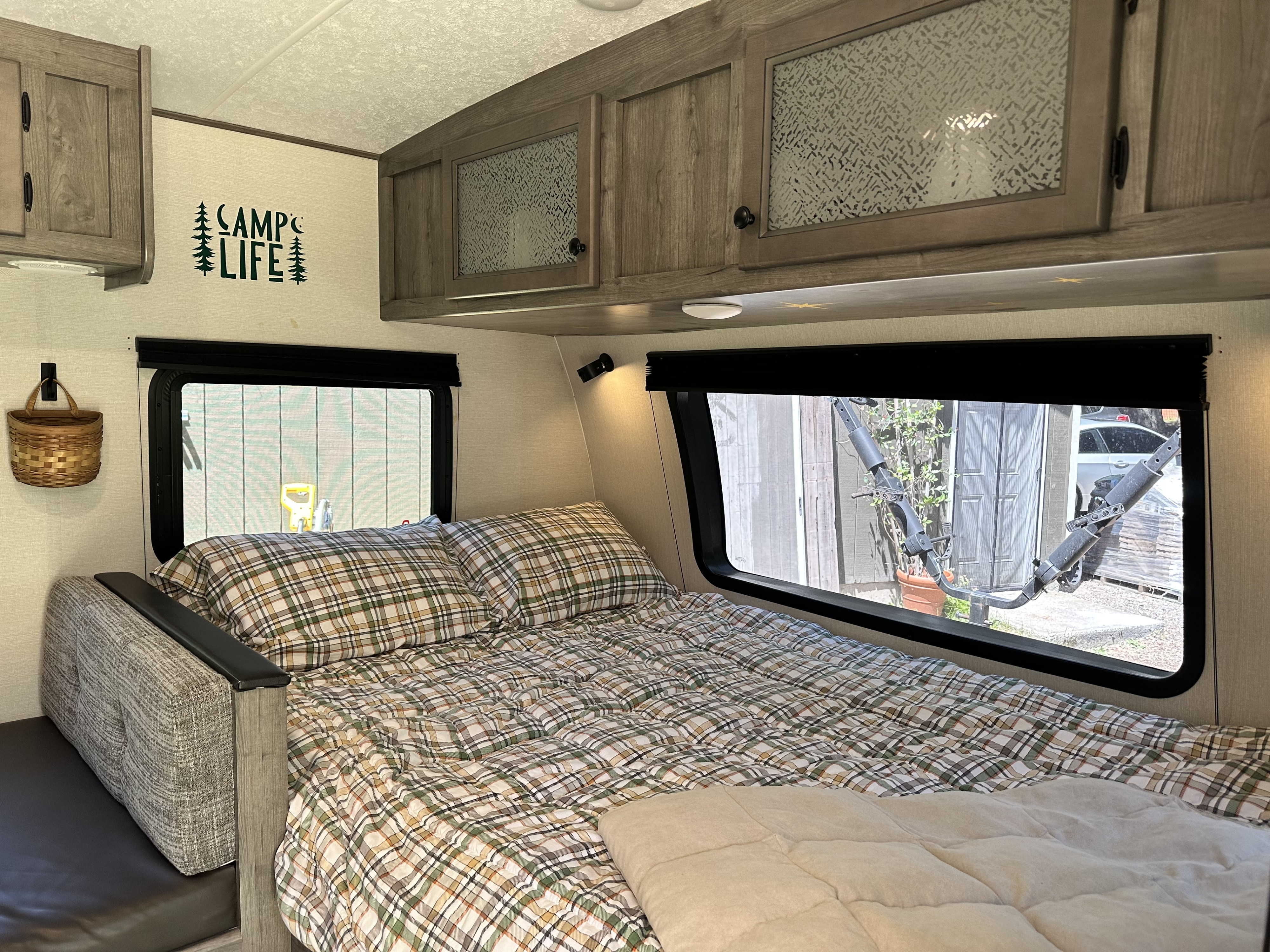 Cozy RV rental bedroom featuring plaid bedding, overhead cabinets, a "CAMP LIFE" decal, and a view outside the window.. Coachmen Apex Nano 2021