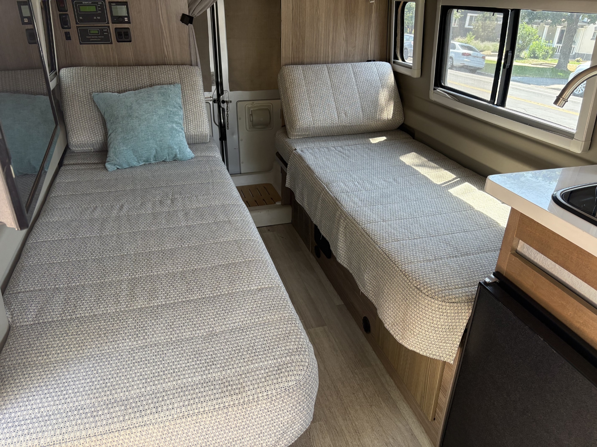 Inside an RV rental: two single beds with light fabric covers, wall controls, and a window showing street views.. Winnebago Travato 2021
