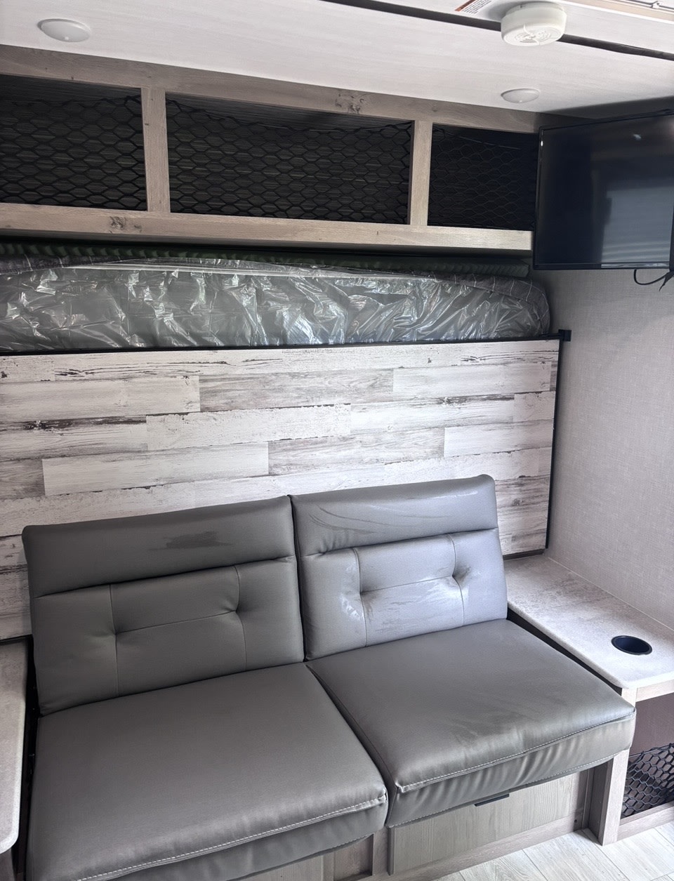 RV rental interior showcasing a gray sofa, an overhead bunk bed (plastic-wrapped), storage, and a wall-mounted TV.. Forest River Surveyor Legend 2022