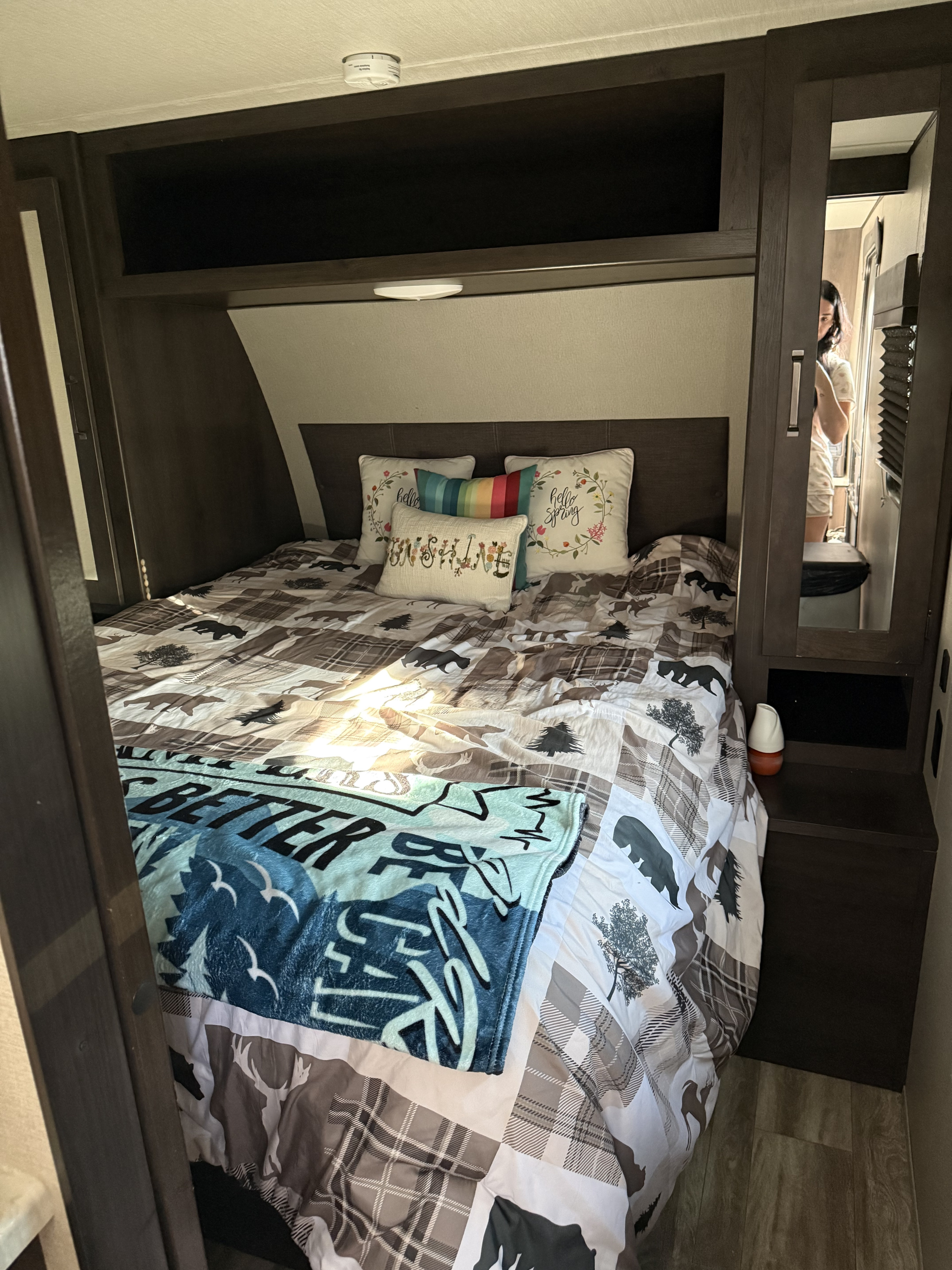 This cozy RV rental bedroom features a bed with plaid and nature-themed bedding, decorative pillows, and storage. A person is reflected in the mirror.. Grand Design Transcend Xplor 2023