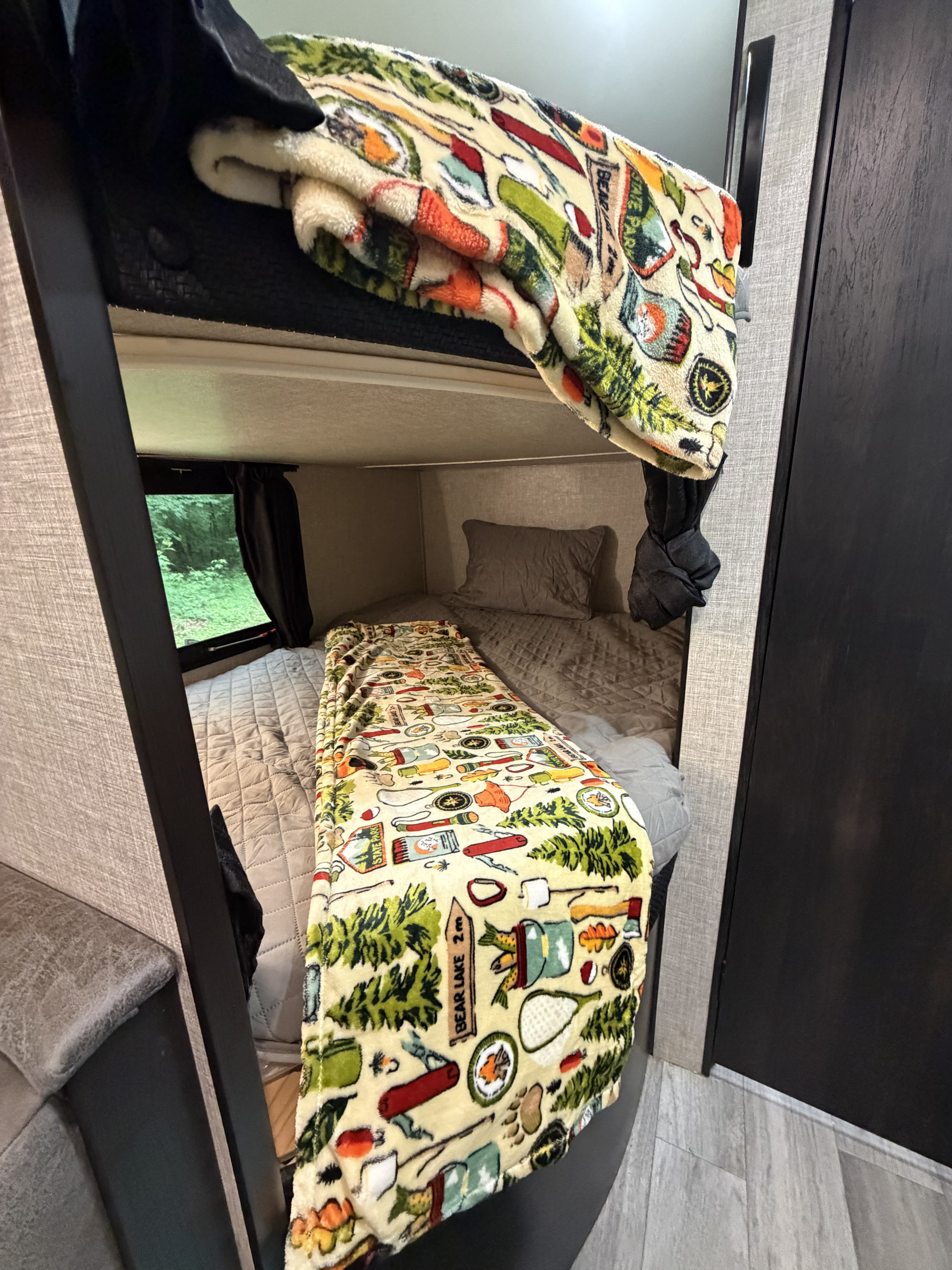 Cozy RV rental bunk beds with camping-themed blankets, ready for adventure. A window shows green trees outside.. Jayco Jay Flight SLX 2022