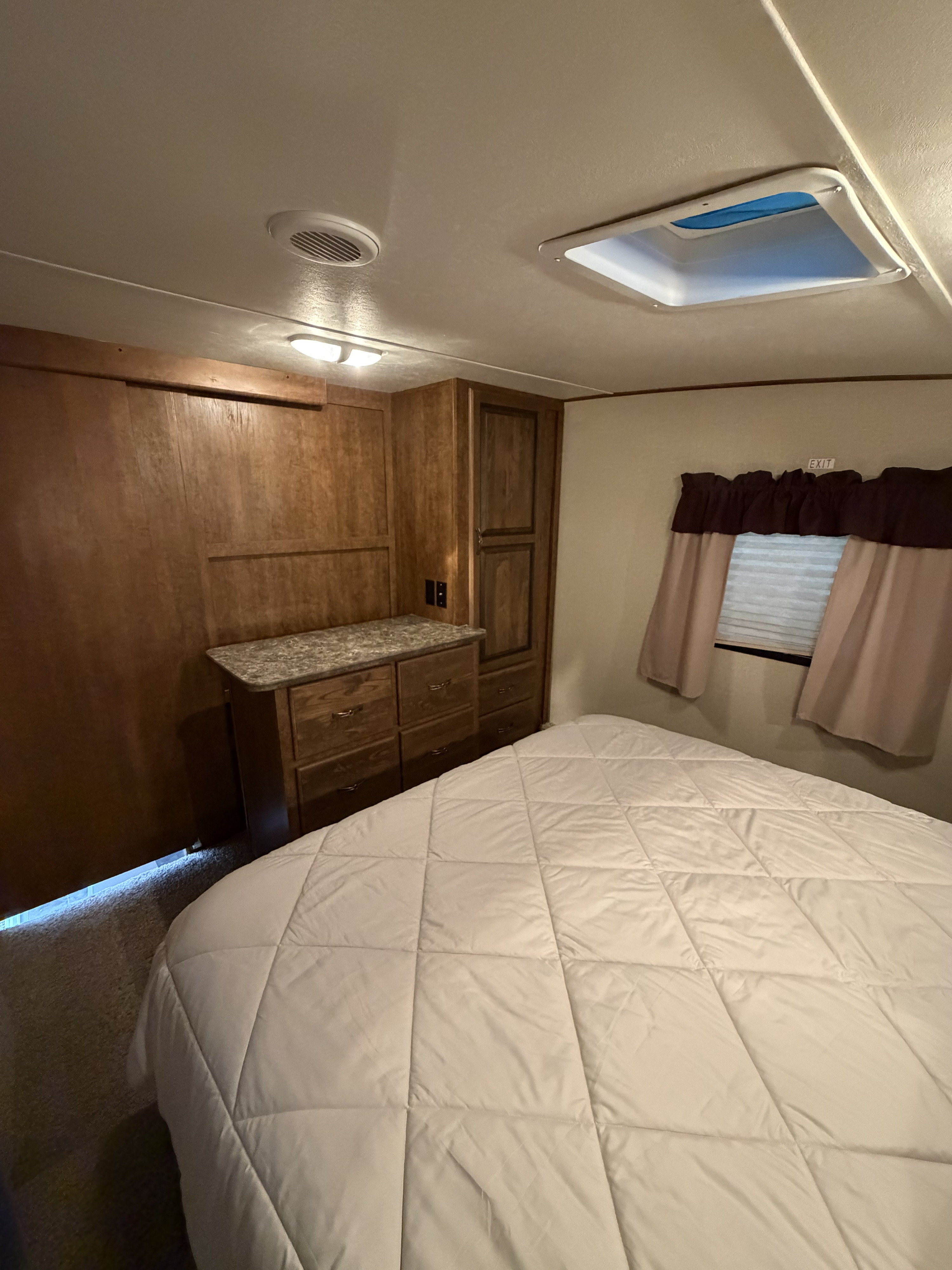 Cozy RV rental bedroom with a white quilted bed, wood cabinetry, dresser, window, ceiling vent, and skylight.. Crossroads RV Rezerve 2016