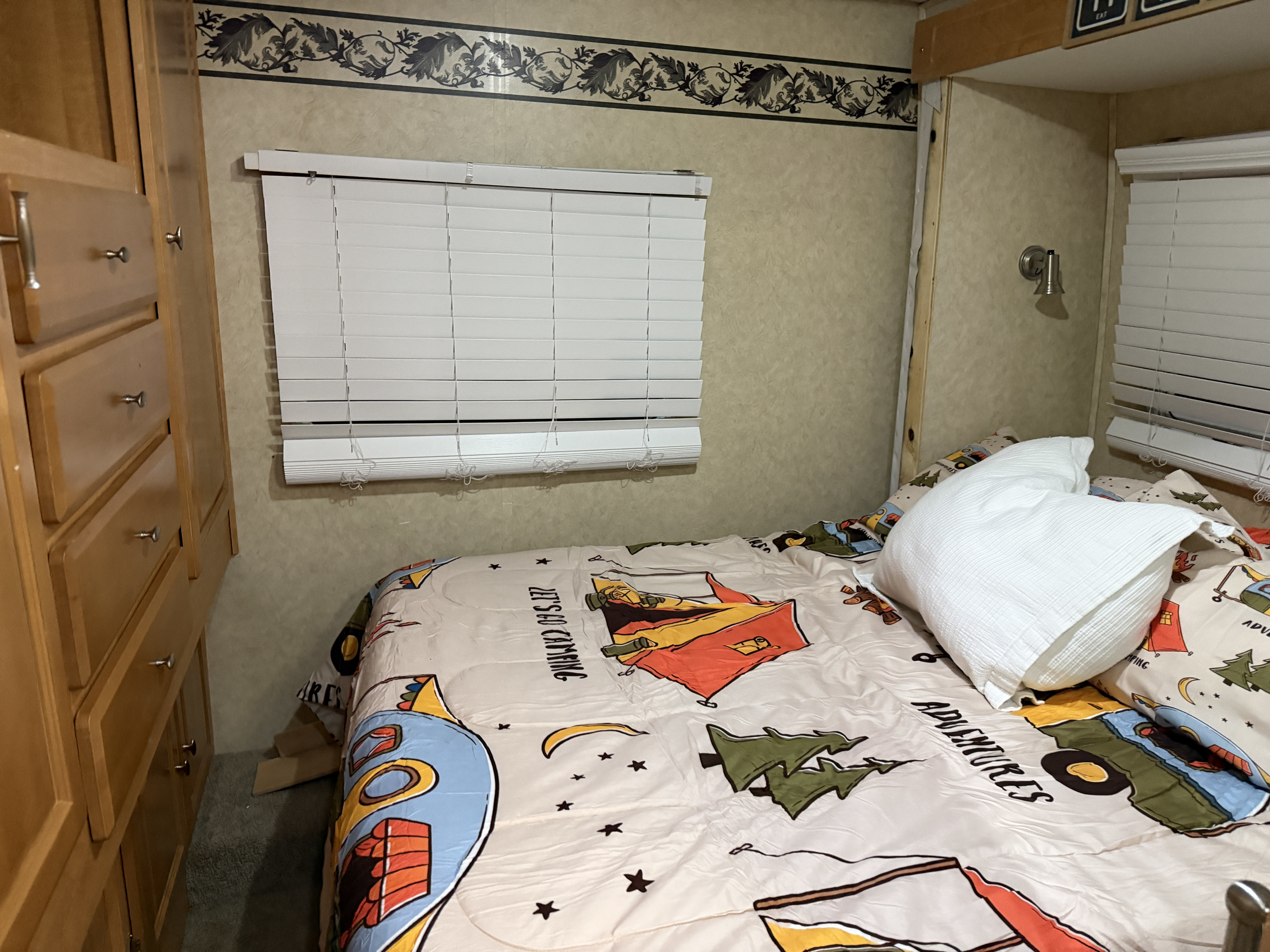 Inside an RV rental: a compact bedroom with a bed, adventure-themed comforter, window blinds, and wood storage.. Lexington Lexington Motorhome 2007