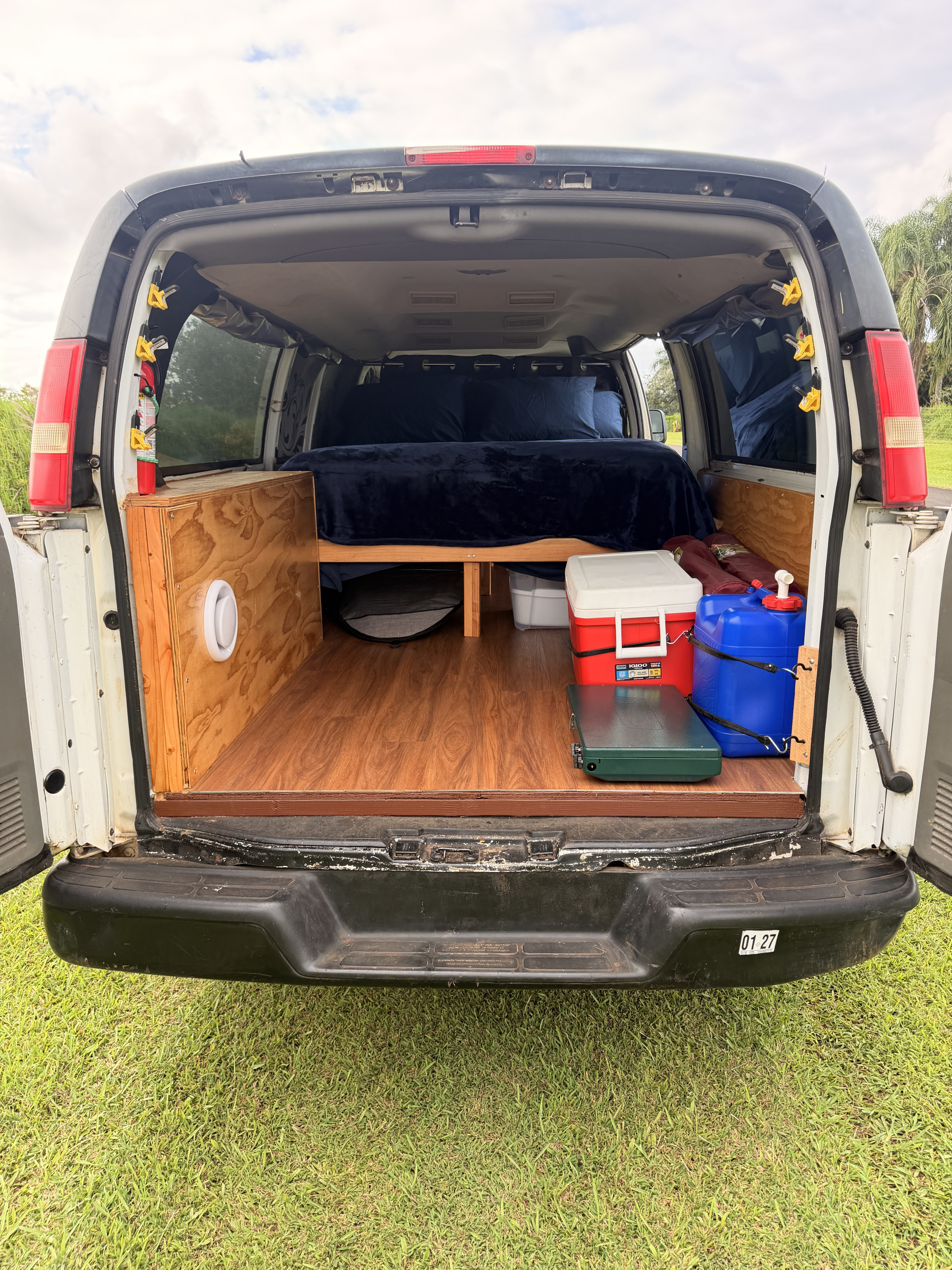 Camper van interior for an rv rental, showing a cozy bed, wooden storage, cooler, and water jugs at the back.. Chevrolet Express 3500 extenses 2009