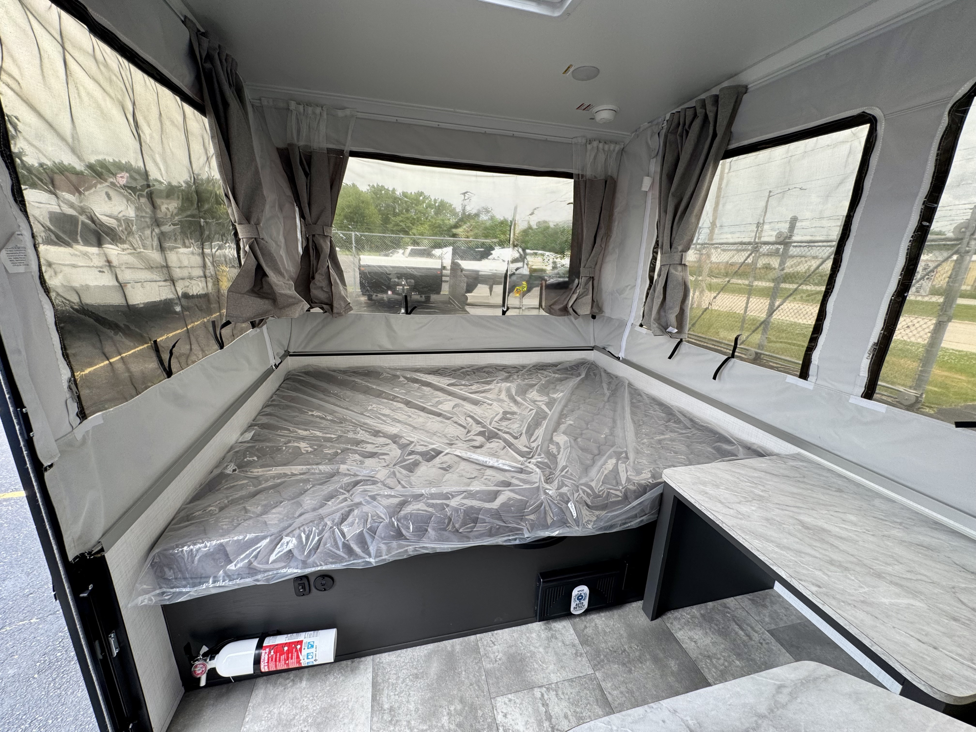 Interior of an RV rental pop-up camper: bed (plastic-covered), side table, windows, fire extinguisher, & ceiling detectors.. Forest River OTG 2025