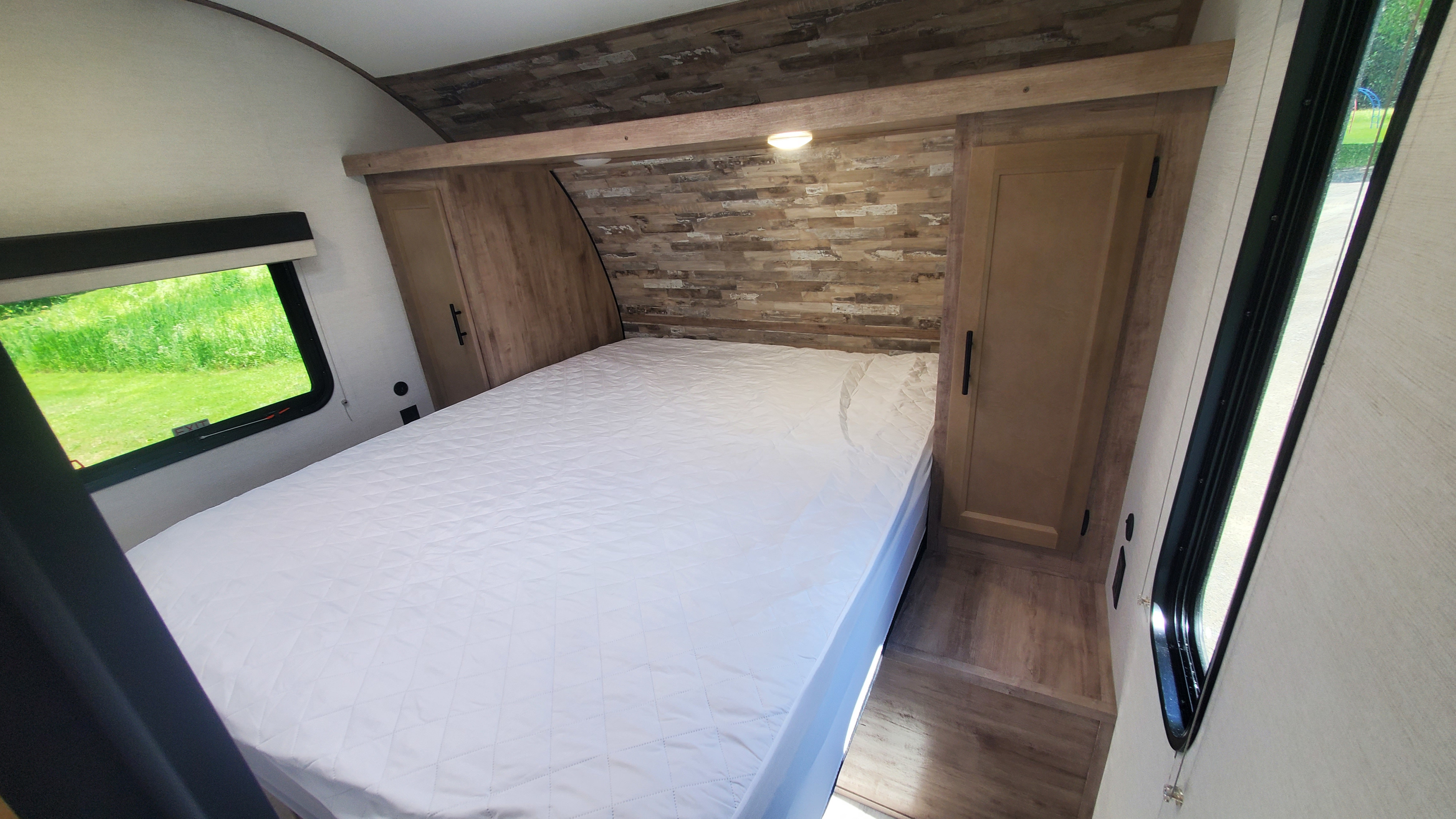 Cozy RV rental bedroom featuring a queen bed, wood-paneled headboard & ceiling, and bright windows with green views.. Gulf Stream Ameri-Lite 2024