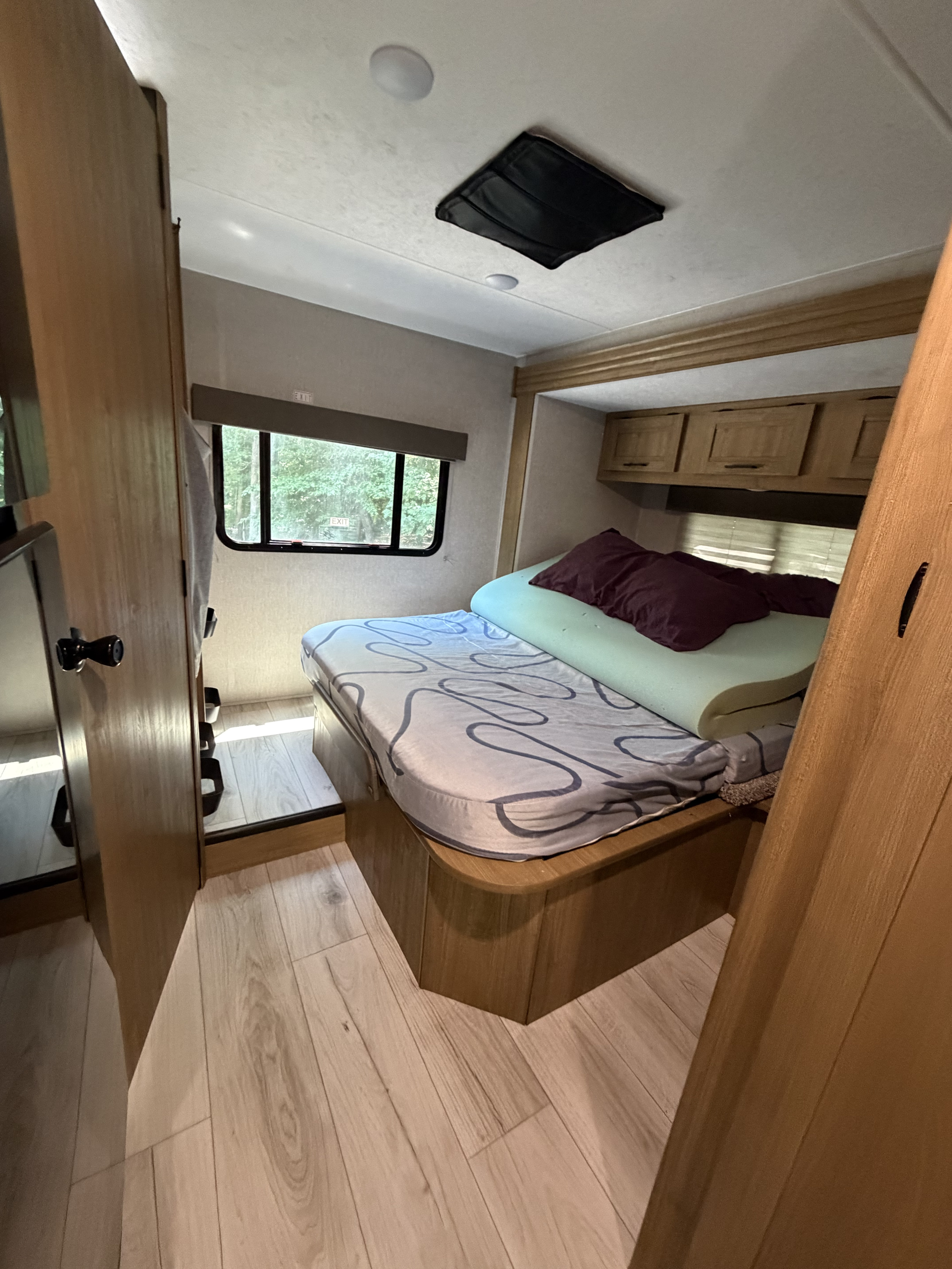 Compact rv rental bedroom: bed with memory foam, wood interior, cabinets, and window view of trees.. Coachman Leprechaun 2025